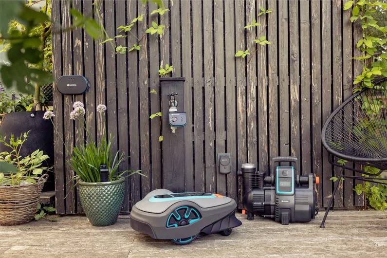 Rasenmähroboter, der auf einer Terrasse in der Nähe von Gartenpflanzen und einem Holzzaun ruht und die automatisierte Gartentechnologie in einer friedlichen Umgebung präsentiert.