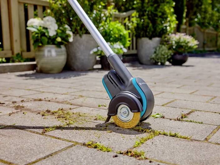 Ein elektrisches Fugenreinigungsgerät entfernt Unkraut zwischen Pflastersteinen auf einem Gehweg, umgeben von Pflanzen in Töpfen.