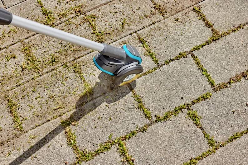Ein Gartenwerkzeug entfernt Moos und Unkraut aus den Zwischenräumen von Pflastersteinen auf einem Gehweg.
