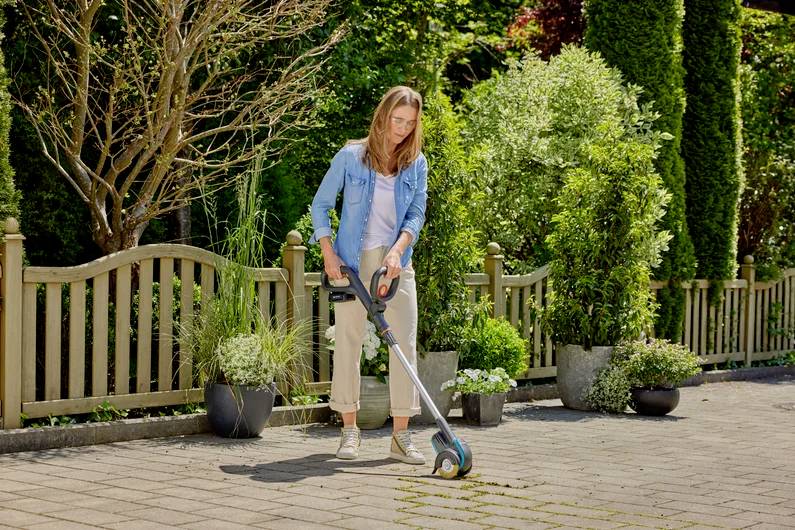 Frau in Freizeitkleidung benutzt einen elektrischen Rasentrimmer, um Unkraut in einem gepflasterten Gartenweg zu entfernen. Hintergrund: dichter Pflanzenwuchs.