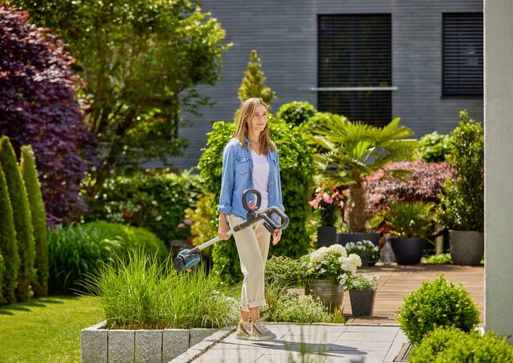 Eine Frau steht mit einem blauen Gartengerät in einem gepflegten Garten vor einem modernen Gebäude. Im Hintergrund sind viele Pflanzen.