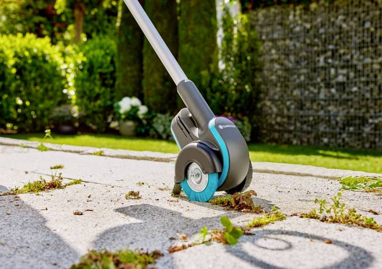 Gartengerät entfernt Unkraut zwischen Steinplatten in einem sonnigen Garten. Im Hintergrund sind Büsche und Rasenschnitt zu sehen.