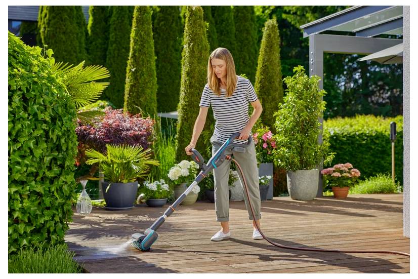 Eine Frau benutzt einen Hochdruckreiniger, um eine Holzterrasse zu reinigen. Üppiges Grün und farbenprächtige Topfpflanzen umgeben sie und schaffen eine friedvolle Outdoor-Umgebung.