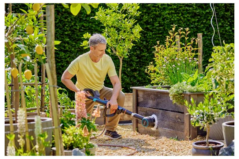 Ein Mann in einem gelben Hemd benutzt einen Gartensprüher, um Pflanzen und erhöhte Holzgartenkästen in einem üppigen, gut gepflegten Garten zu bewässern.
