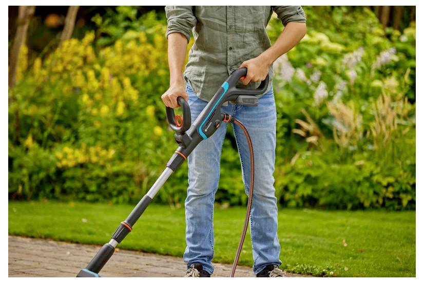 Eine Person in Jeans und einem langärmligen Hemd benutzt einen elektrischen Trimmer auf einem sauber geschnittenen Rasen, mit einem Garten im Hintergrund.