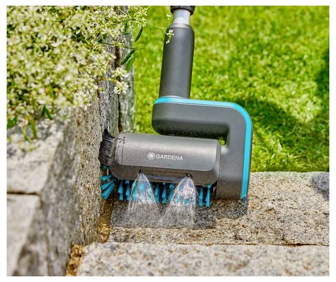 Ein Gartengerät mit blauen Borsten wird auf einer Steinoberfläche verwendet, wobei Wasser versprüht wird, um Moos und Ablagerungen zwischen Pflastersteinen und einer Mauer zu reinigen.