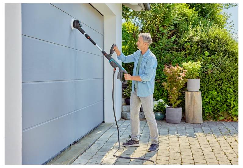 Ein Mann in legerer Kleidung benutzt ein Elektrowerkzeug, um ein Garagentor im Freien zu reinigen, umgeben von Grünpflanzen und Gartendekoration.