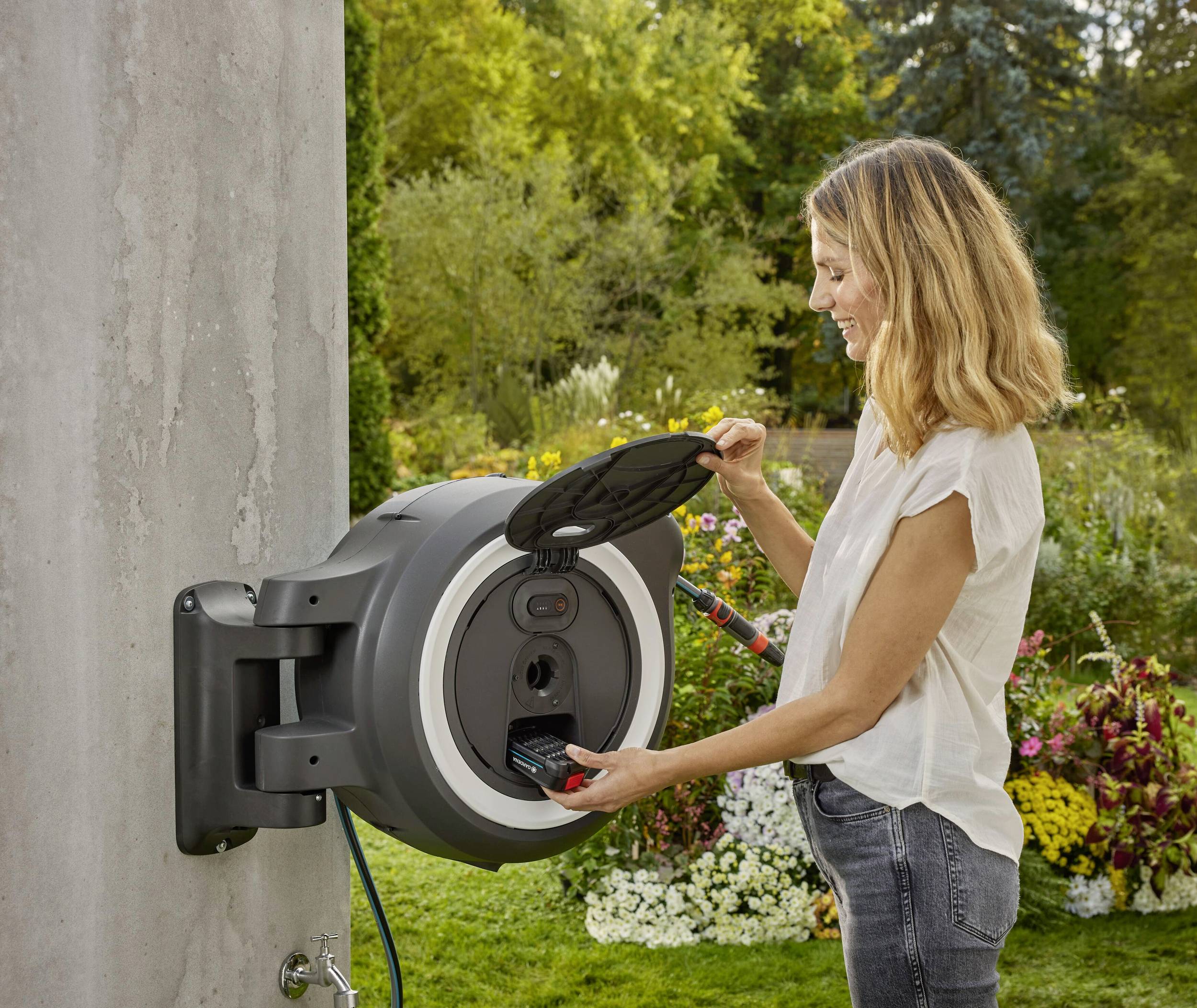 Eine Frau benutzt eine runde Wandbox im Freien zum Aufrollen eines Gartenschlauchs, umgeben von blühenden Pflanzen und Bäumen.
