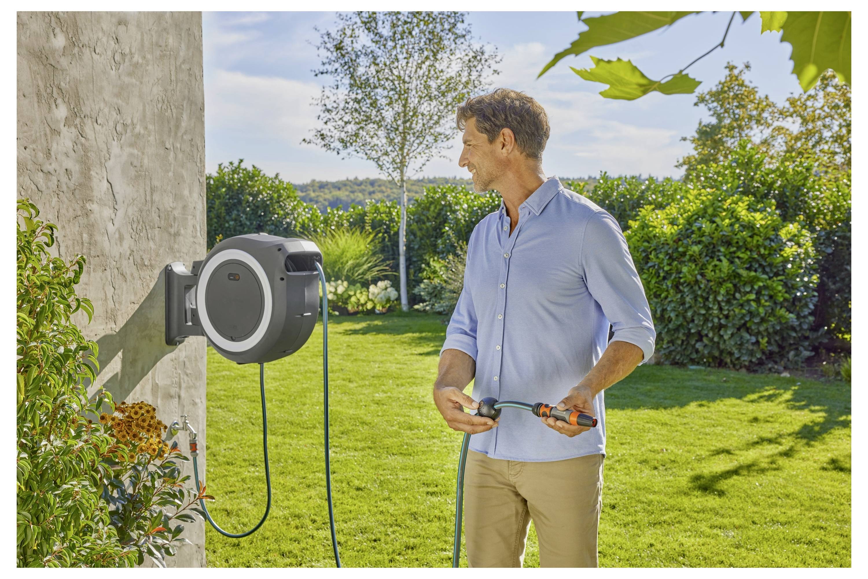 Eine Person in einem hellblauen Hemd steht in einem sonnigen Garten und hält einen Gartenschlauch an einer wandmontierten Schlauchrolle, umgeben von grünen Sträuchern.