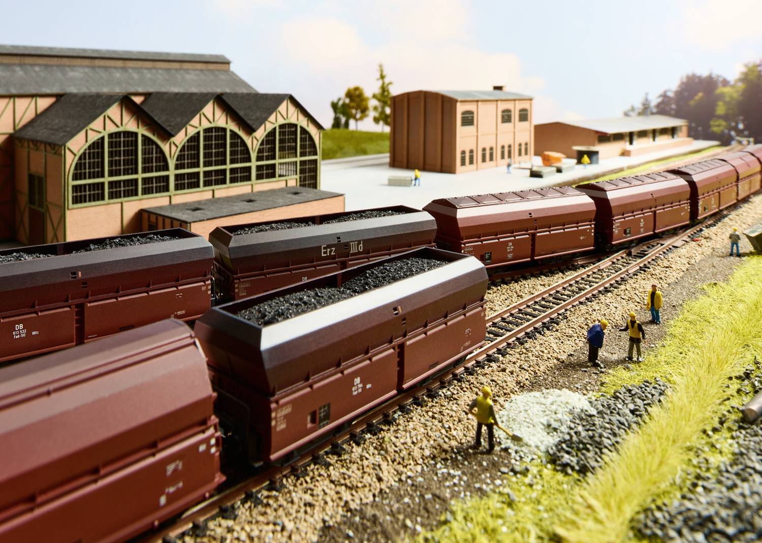 Modelleisenbahn-Szene mit mehreren Güterwaggons voller Kohle auf Schienen vor einem Bahnhofsgebäude, umgeben von Miniaturmenschen.