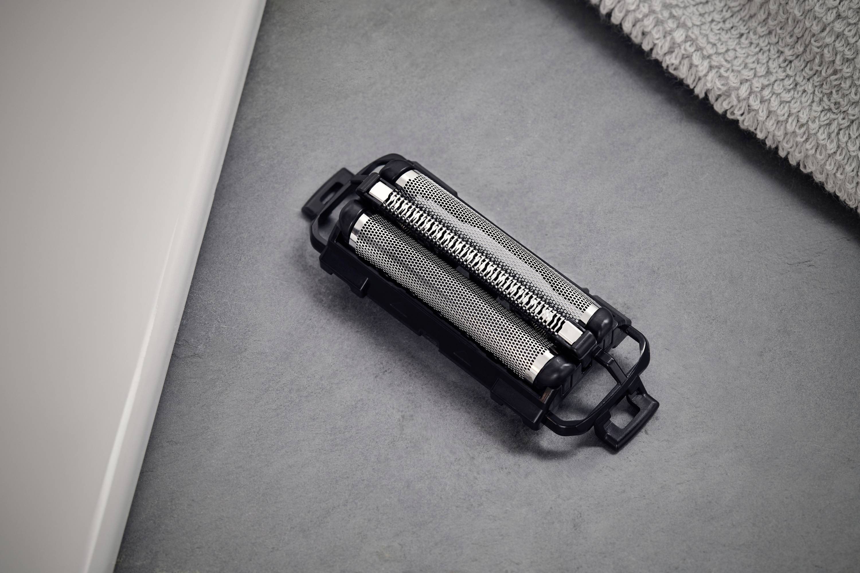 Rasiererkopf auf grauem Hintergrund, neben einem Handtuch und einer Kante eines weißen Waschbeckens.