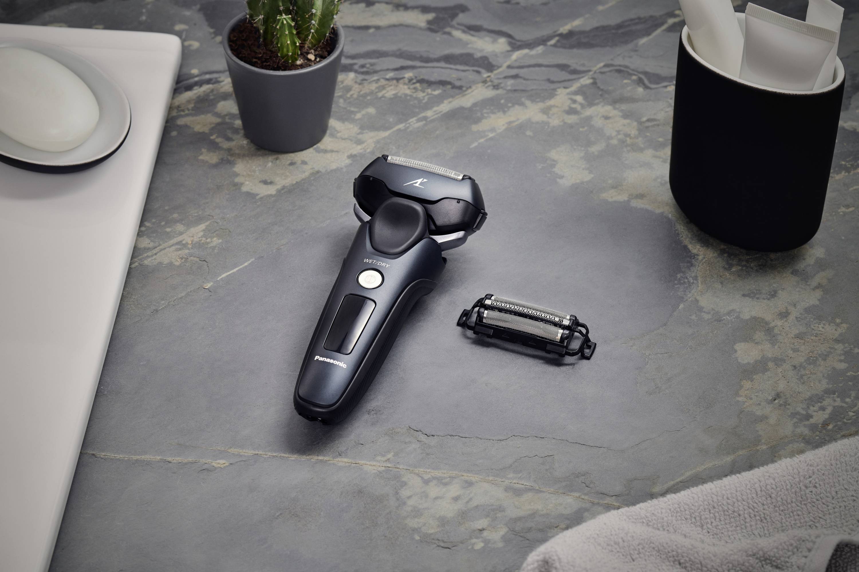 Elektrorasierer mit zusätzlichem Scherkopf auf grauer Oberfläche, neben Pflanze und Bechern, vermitteln Badezimmer- oder Pflegeszenario.