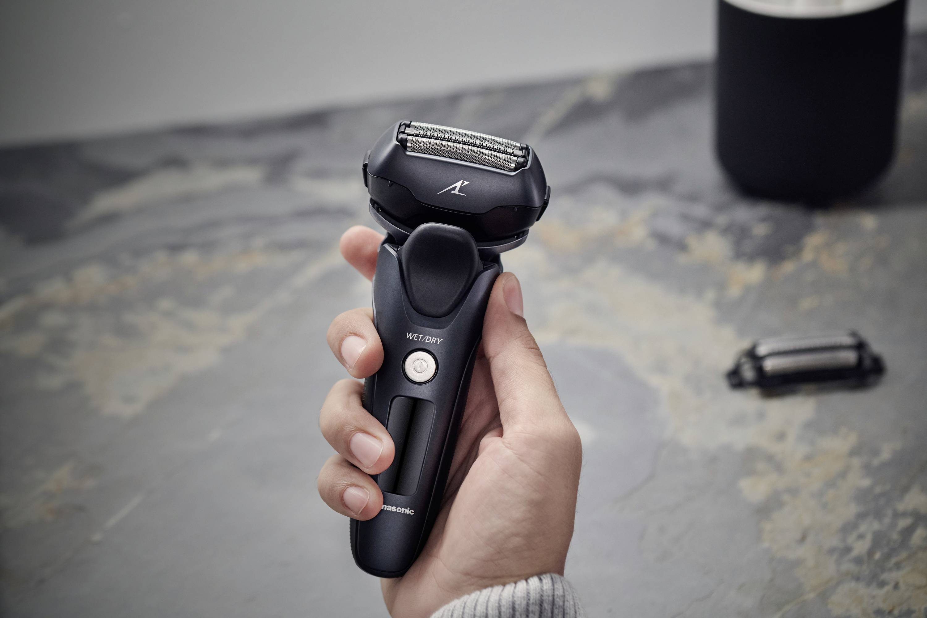 Eine Hand hält einen modernen elektrischen Rasierer über einer grauen Oberfläche. Rechts im Hintergrund liegt ein abgenommenes Rasiererteil.