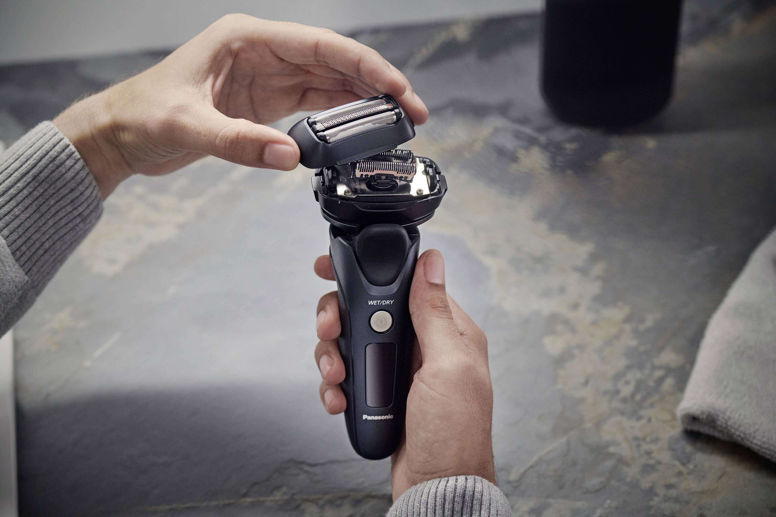 Eine Person hält einen elektrischen Rasierer und öffnet den Scherkopf. Auf einem grauen Tisch im Hintergrund liegt ein Handtuch.