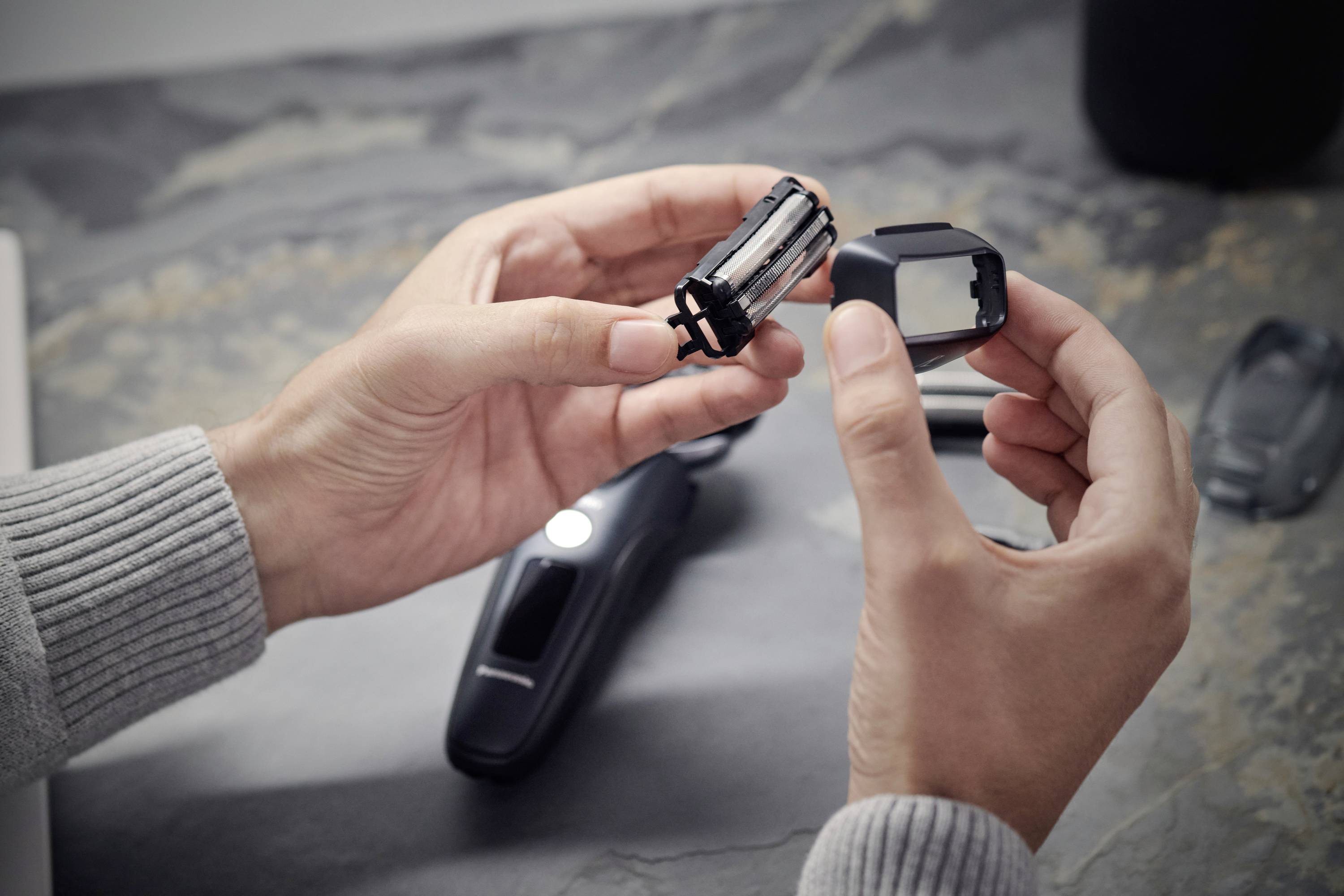 Eine Person hält Ersatzteile eines elektrischen Rasierers in den Händen, auf einem Tisch liegt der Rasierer im Hintergrund.