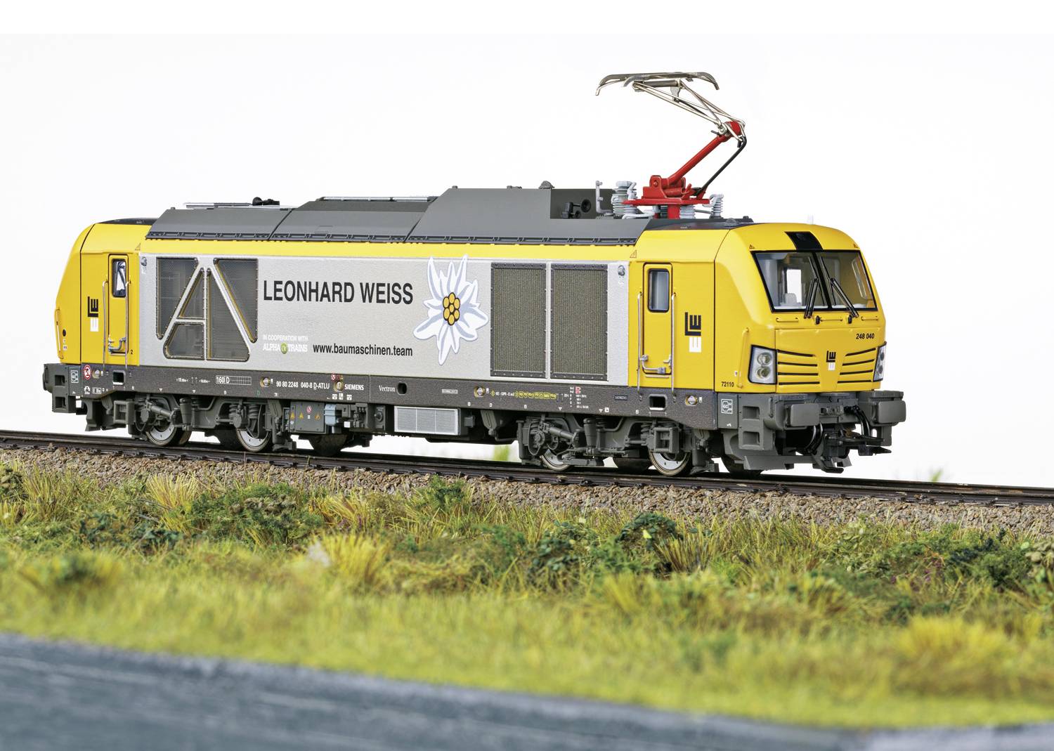 Gelbe Lokomotive mit grauem Streifen und 'Leonhard Weiss' Logo fährt auf einer Schiene, Umgebung mit Gras und Bahntrasse.