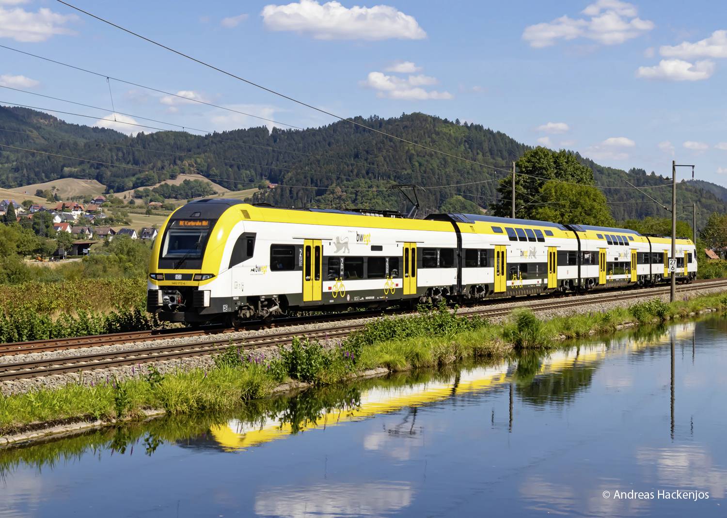 Gelb-weißer Zug fährt entlang eines Sees in ländlicher Landschaft mit Hügeln und Blauem Himmel im Hintergrund.