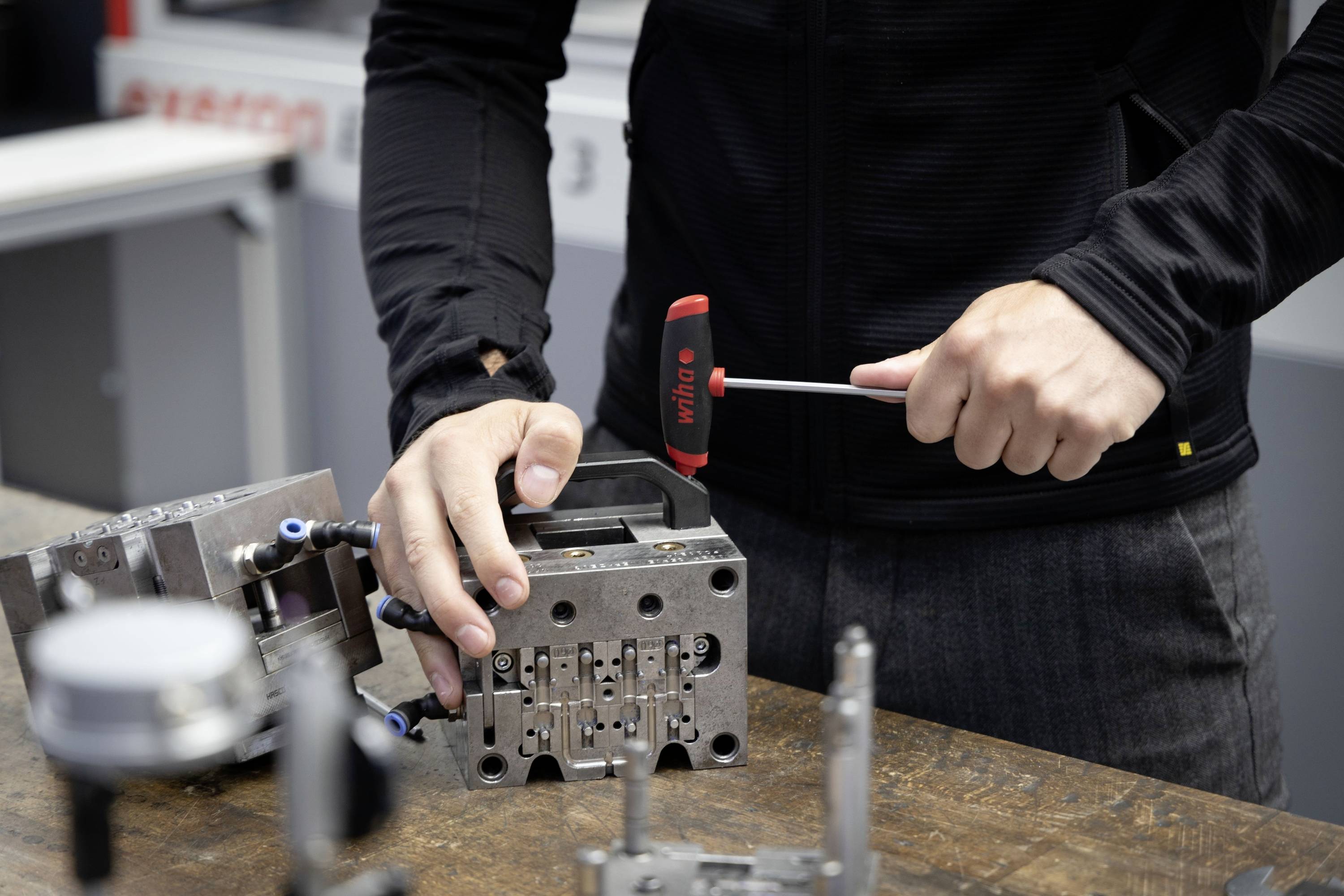 Eine Person in schwarzer Kleidung arbeitet mit einem roten Hammer an einer Metallform auf einem Holztisch in einer Werkstattumgebung.