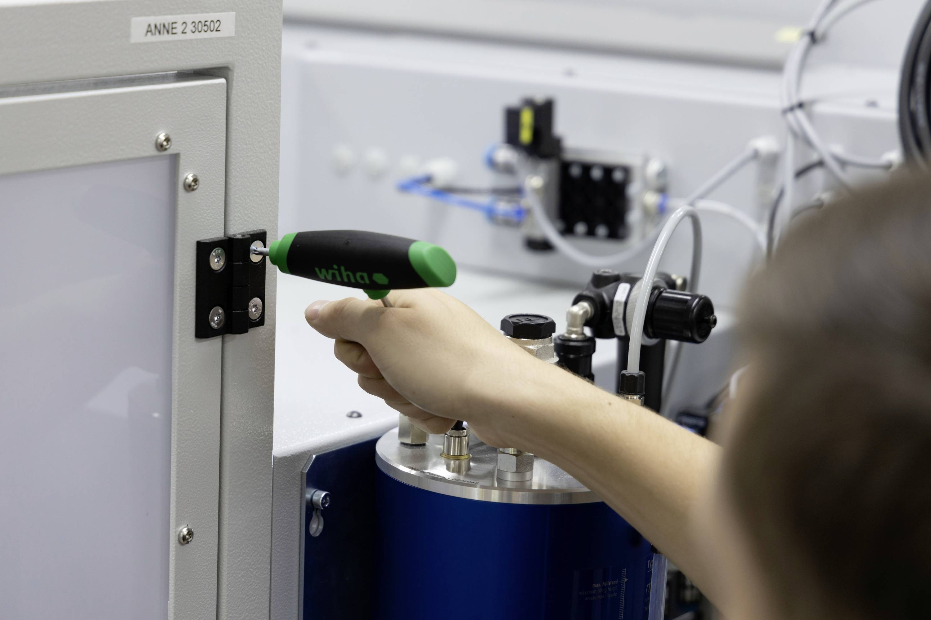 Eine Person montiert eine Schranktür mit einem Schraubendreher. Im Hintergrund sind industrielle Maschinen und Kabel zu sehen.