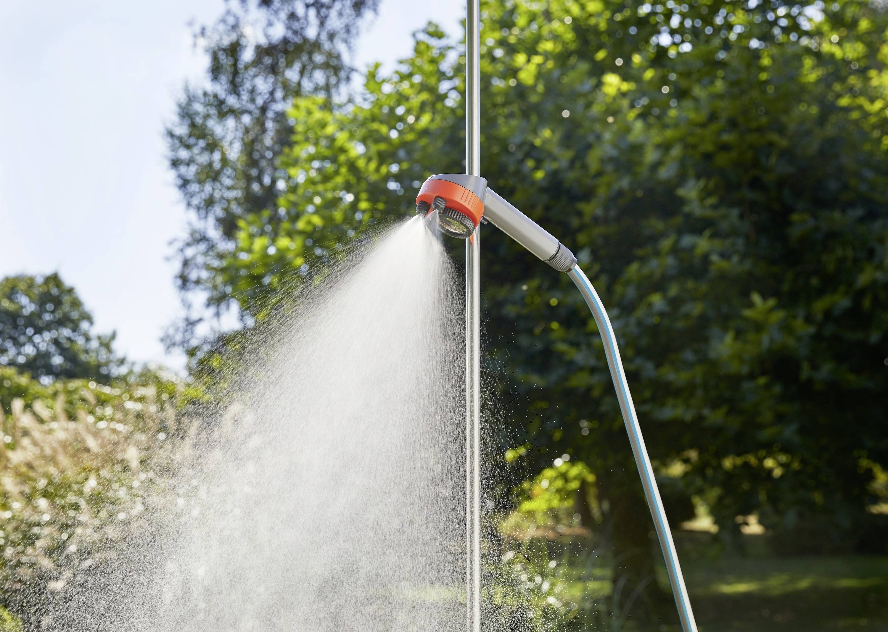 Gartendusche im Freien, die Wasser versprüht, positioniert in einer grünen Hofumgebung mit Bäumen und Sonnenlicht im Hintergrund.