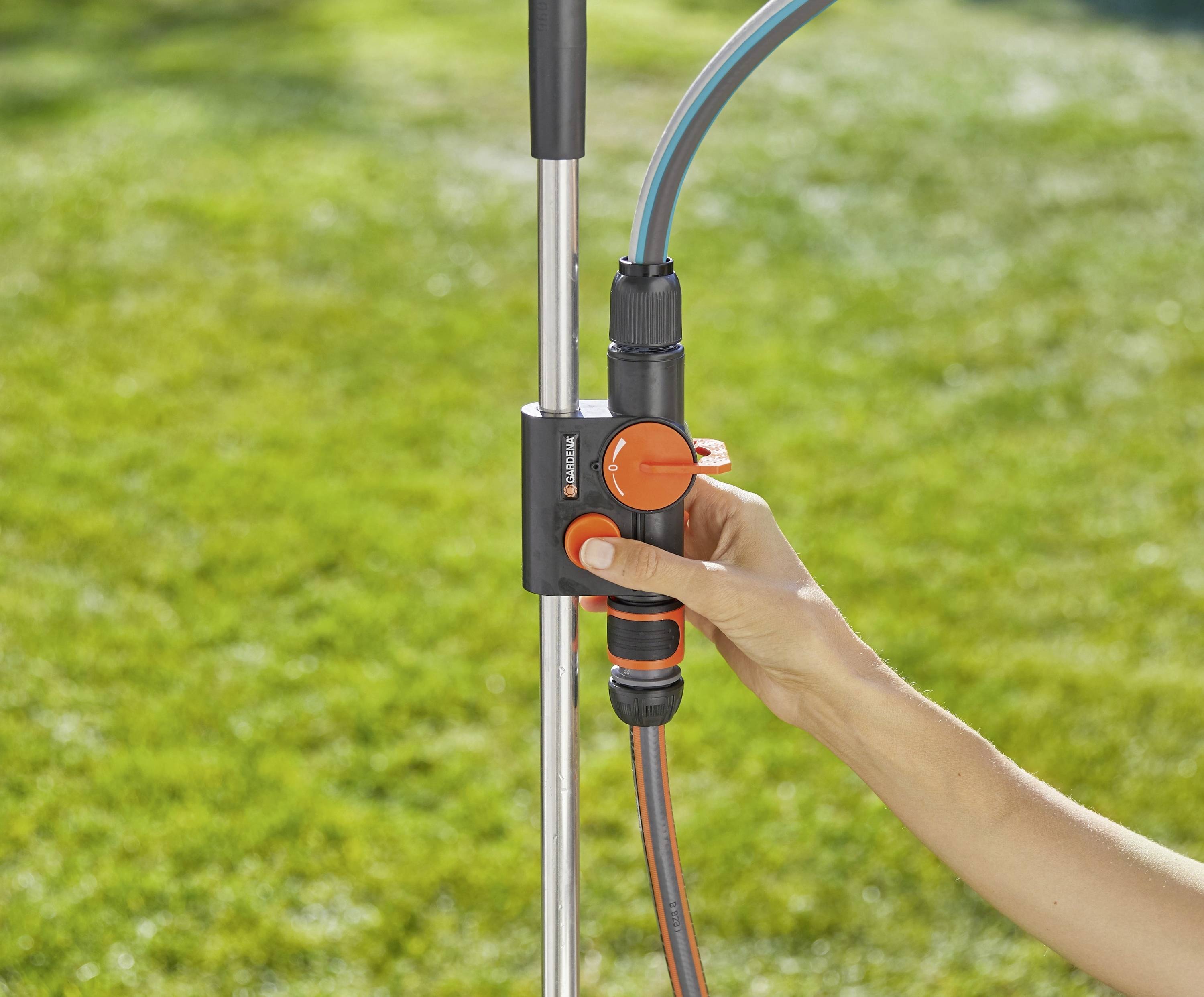 Eine Hand justiert ein schwarzes und orangefarbenes Gartenventil zur Wasserdurchflusskontrolle, das an einem Metallpfosten befestigt ist, mit einem Rasen im Hintergrund.