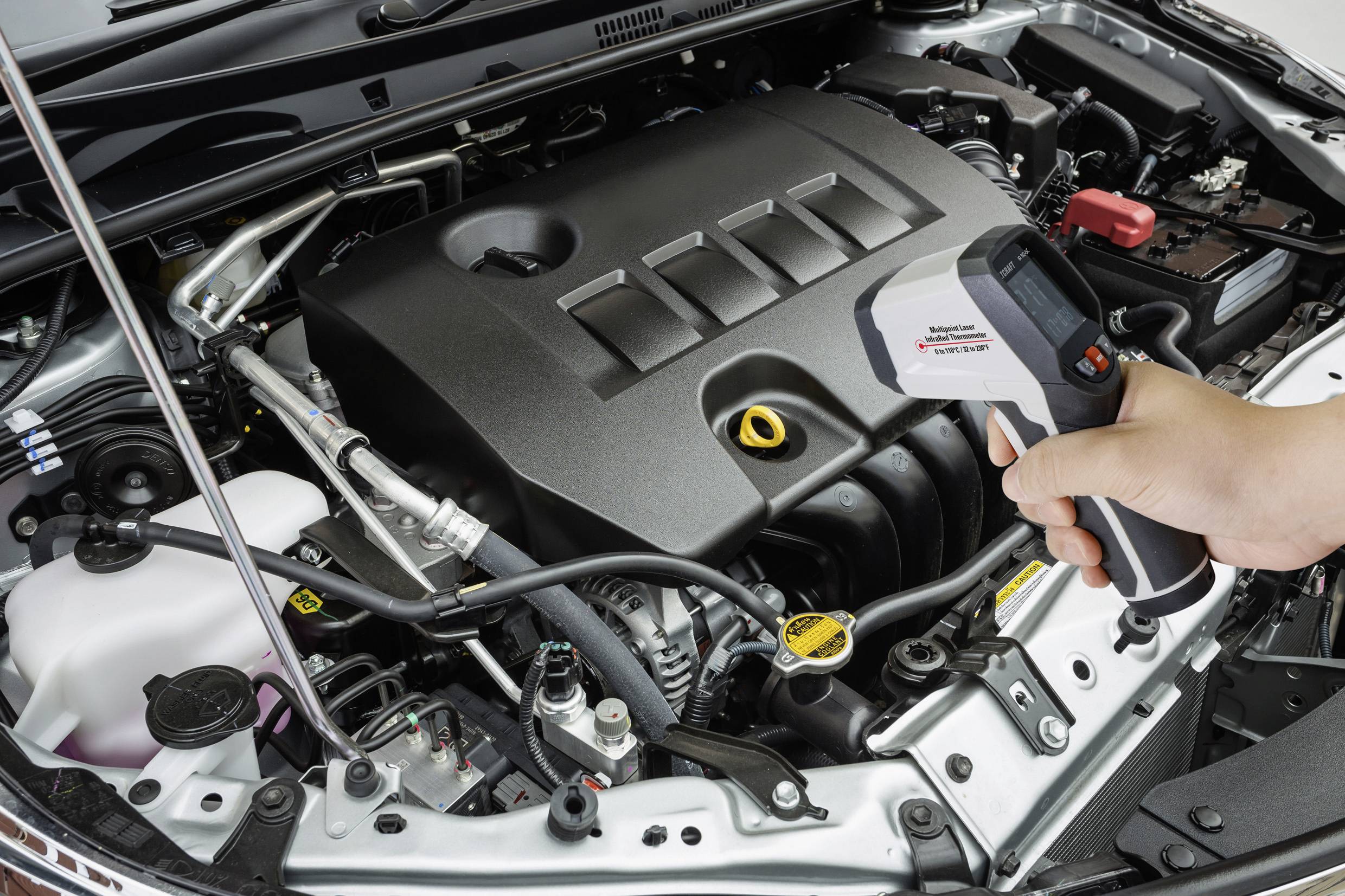 Person hält ein digitaler Temperaturmesser über geöffnete Motorhaube eines Autos. Messgerät zeigt 35,2 Grad Celsius.