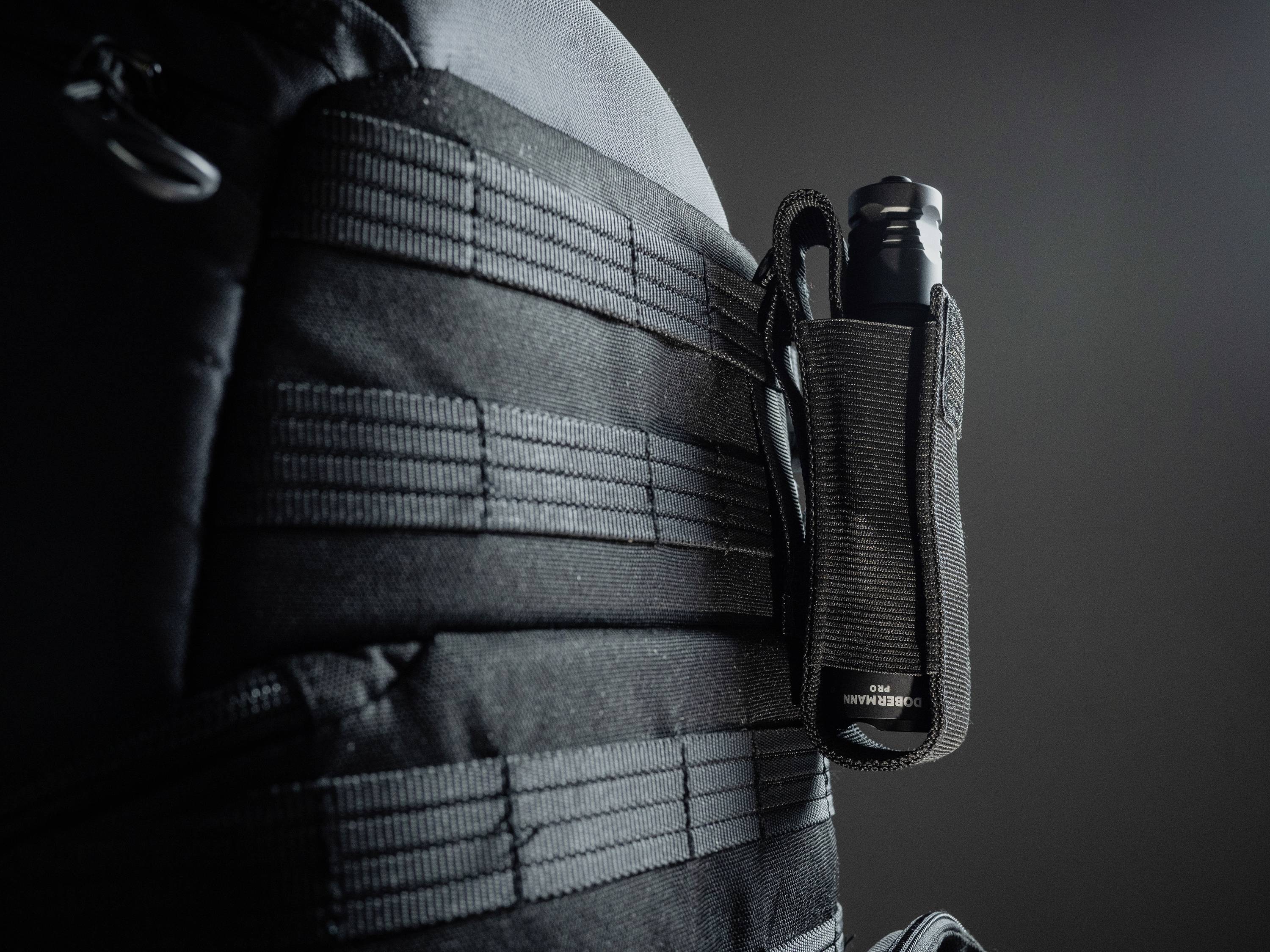 Ein taktischer Rucksack mit Schlaufen und einer befestigten schwarzen Taschenlampe an der Seite vor einem dunklen Hintergrund.