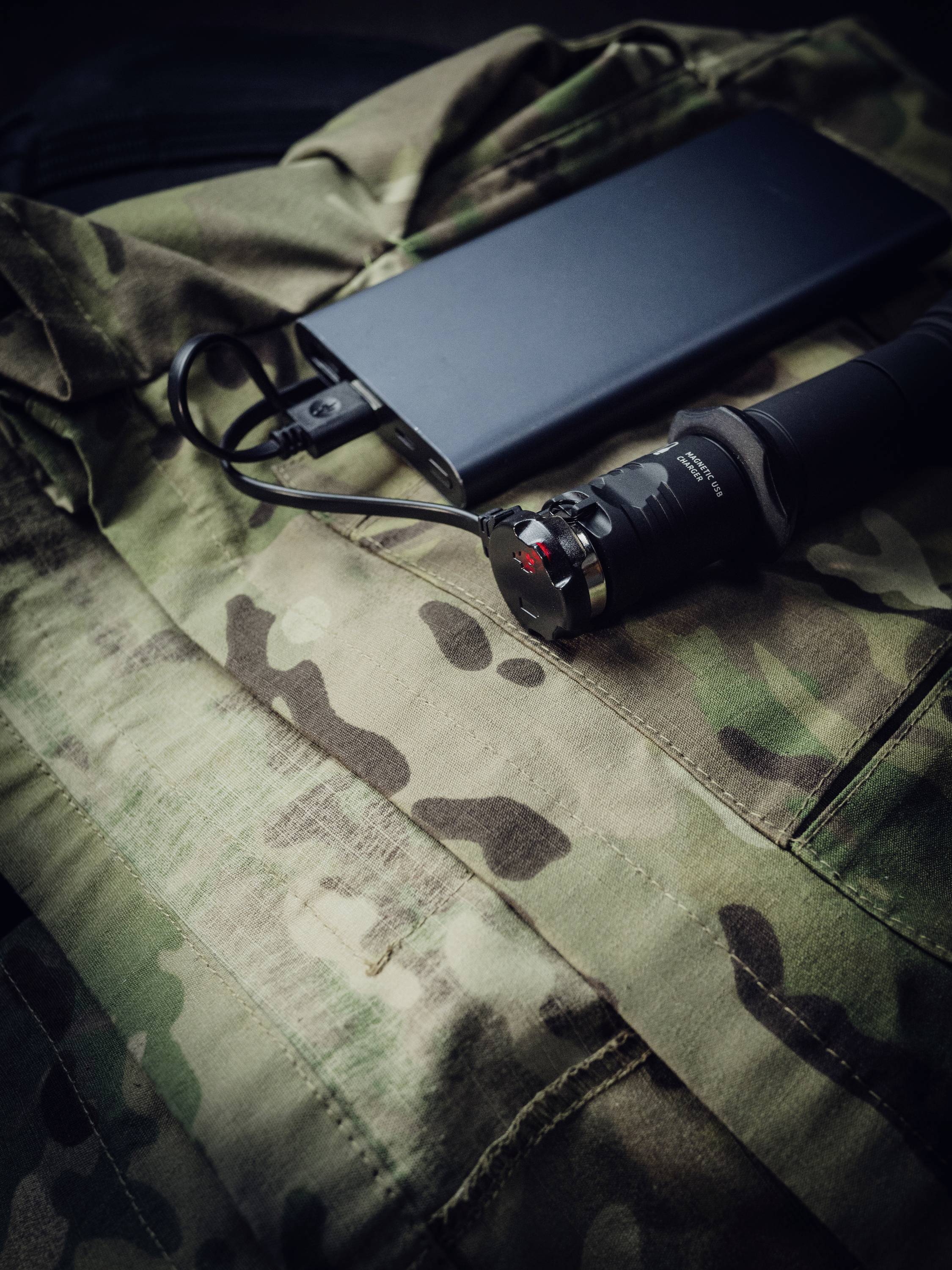 Taktische Ausrüstung auf einer Tarnjacke: eine Taschenlampe neben einer Powerbank, beide verbunden durch ein Ladekabel.