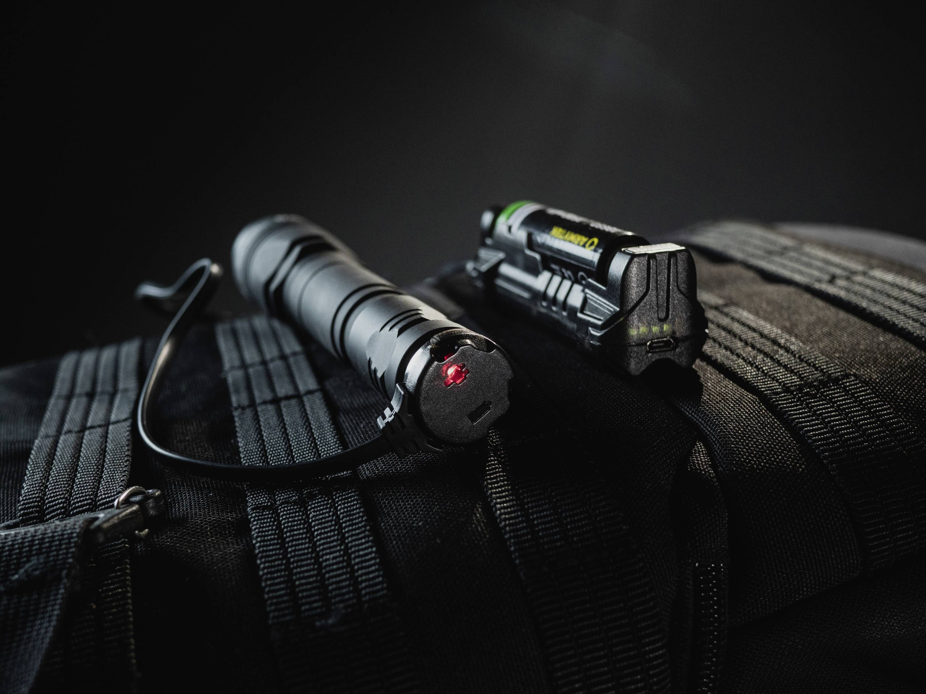 Taschenlampe mit rotem Licht auf schwarzem Rucksack, daneben eine Lithium-Batterie. Dunkler Hintergrund, Fokus auf Outdoor-Ausrüstung.