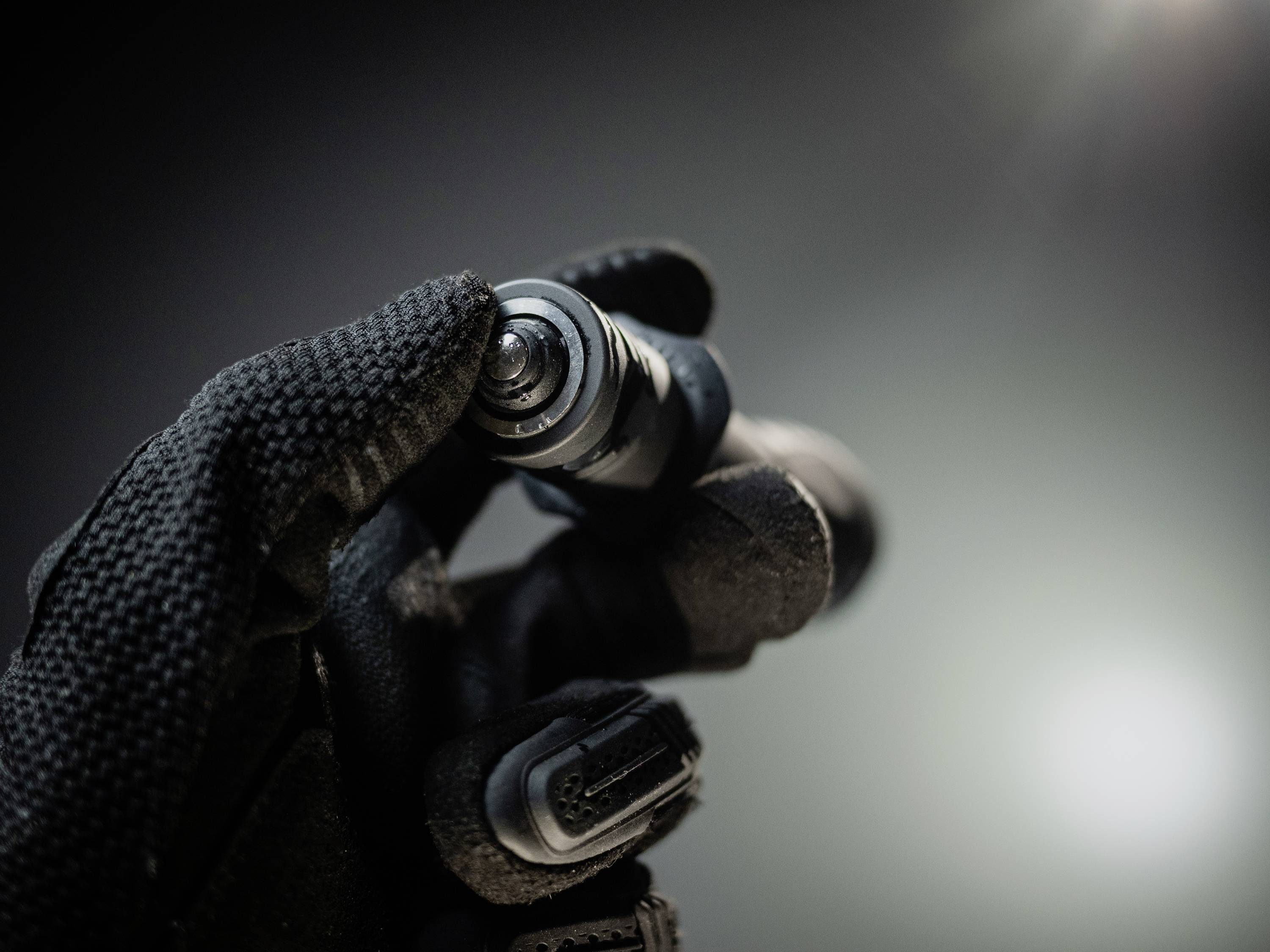 Eine behandschuhte Hand hält ein kleines technisches Gerät in dunkler Umgebung. Es scheint sich um ein elektronisches Bauteil zu handeln.