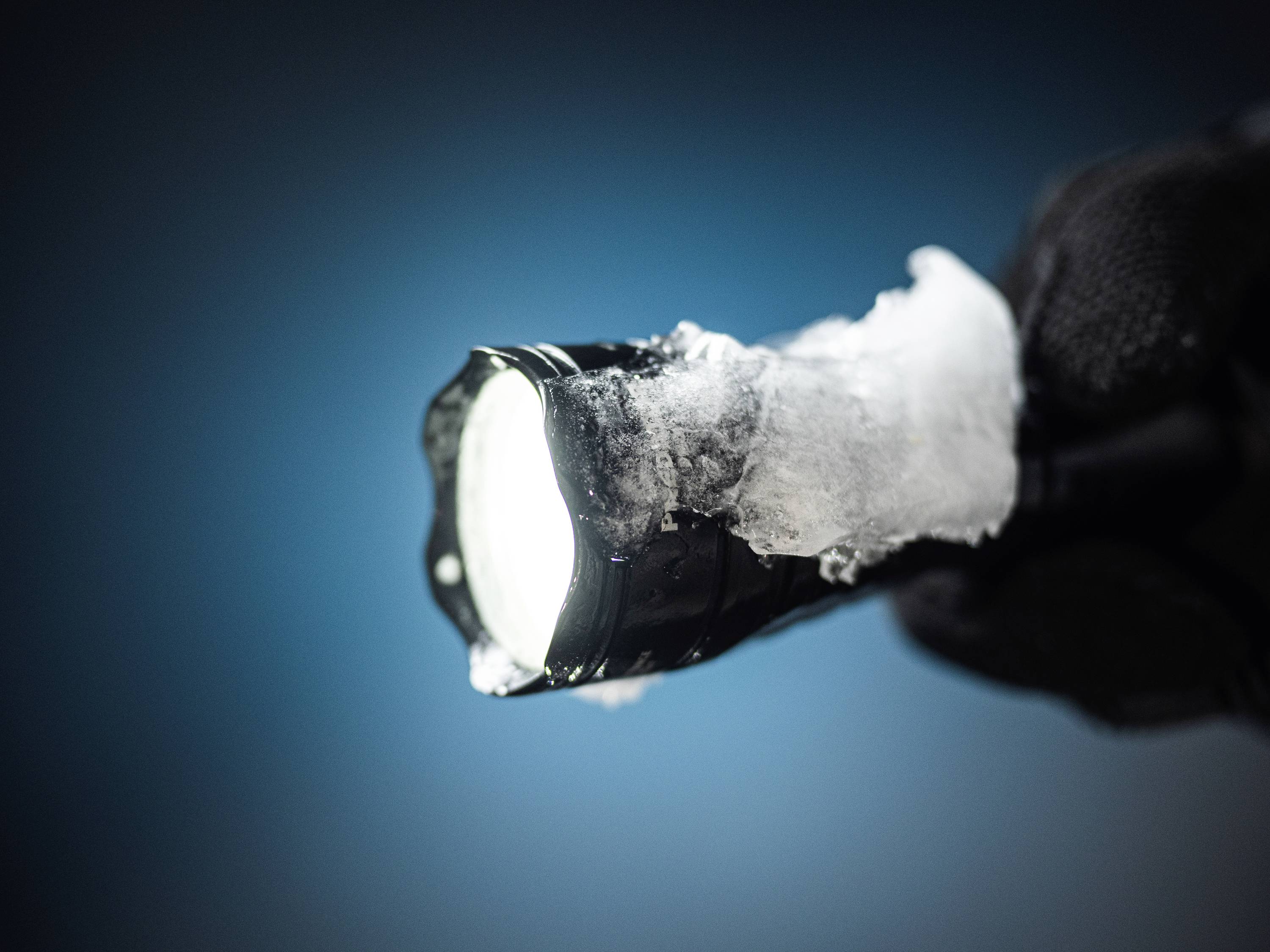 Hand mit Handschuh hält eine eingeschaltete Taschenlampe, die mit Schnee bedeckt ist.