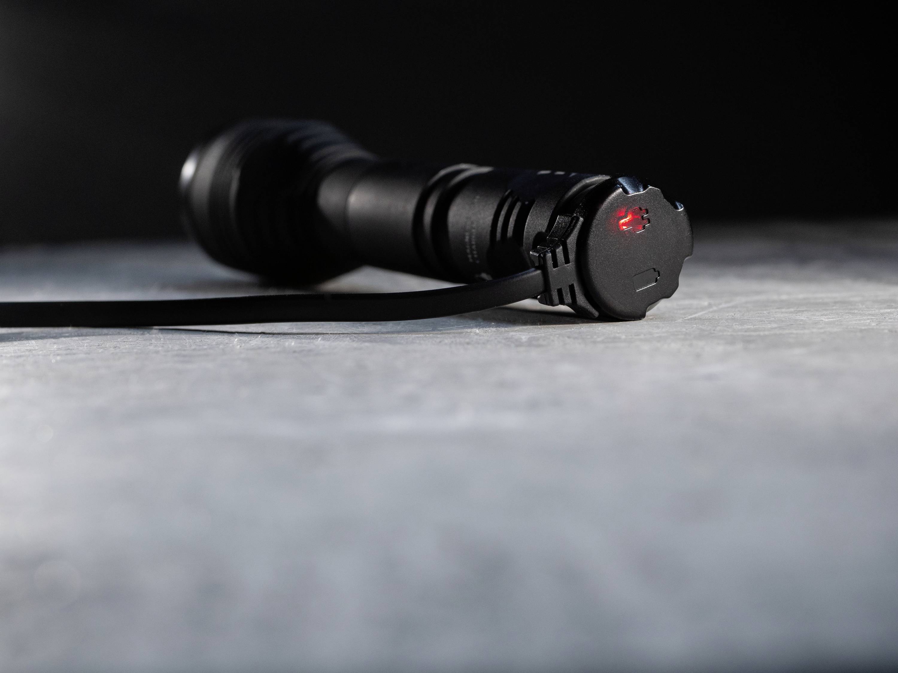 Eine schwarze Taschenlampe liegt horizontal auf einer grauen Oberfläche. Ein schwaches rotes Licht leuchtet am Ende.