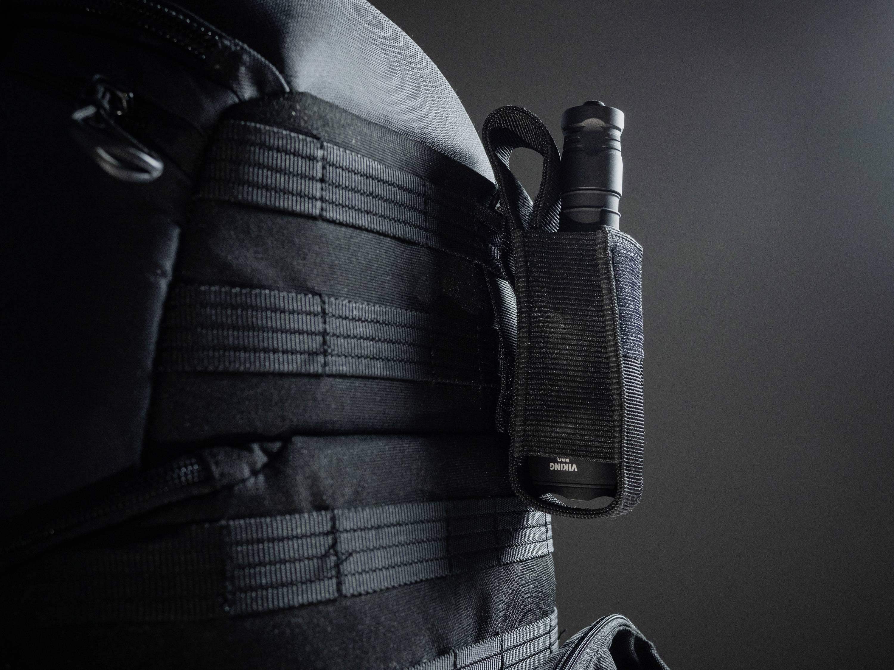 Ein schwarzer Rucksack mit einer Taschenlampe in einer Halterung an der Seite, auf einem dunklen Hintergrund.