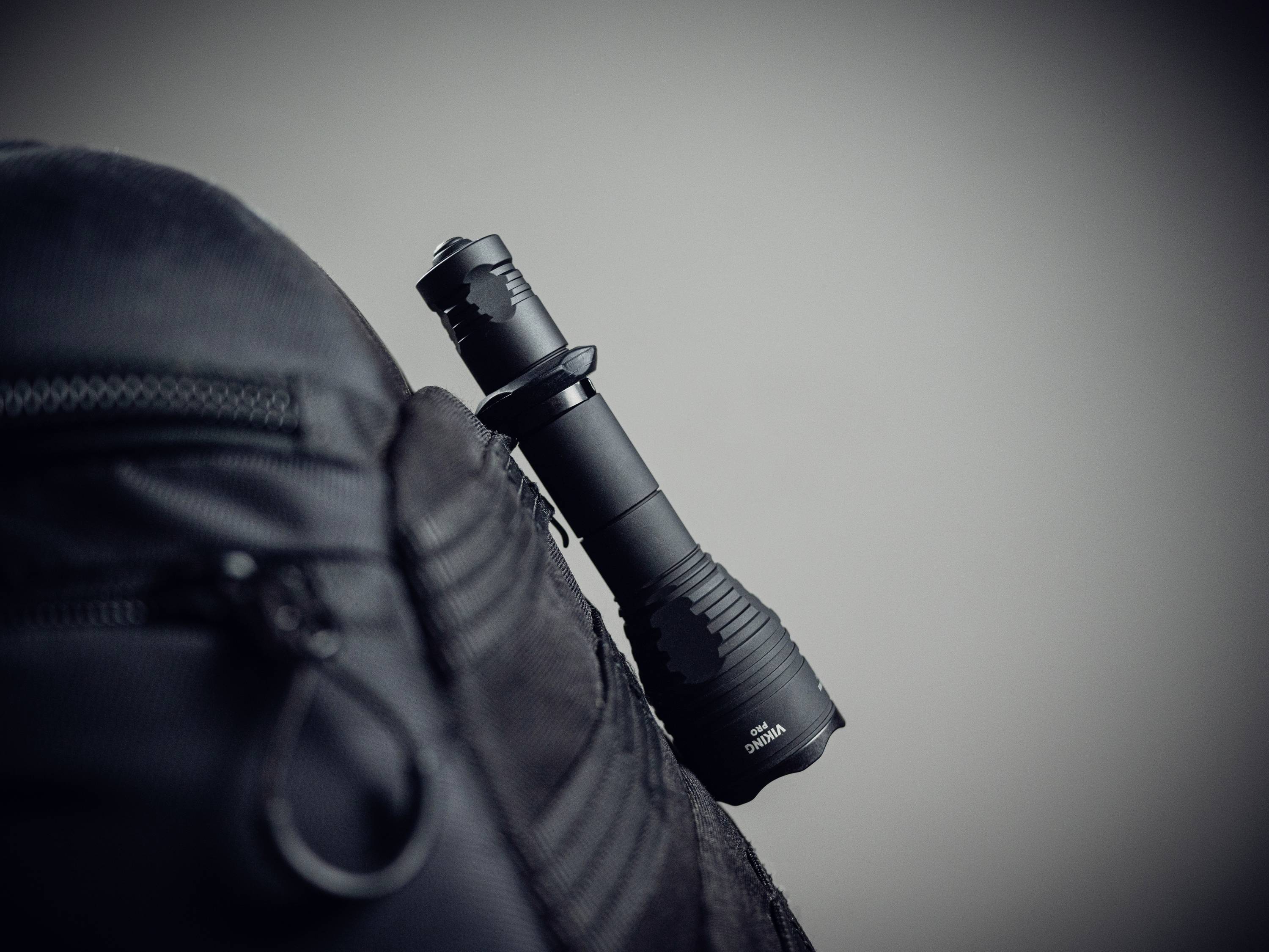 Taschenlampe in schwarz steckt seitlich in einer Tasche eines schwarzen Rucksacks. Hintergrund ist unscharf und dunkelgrau.