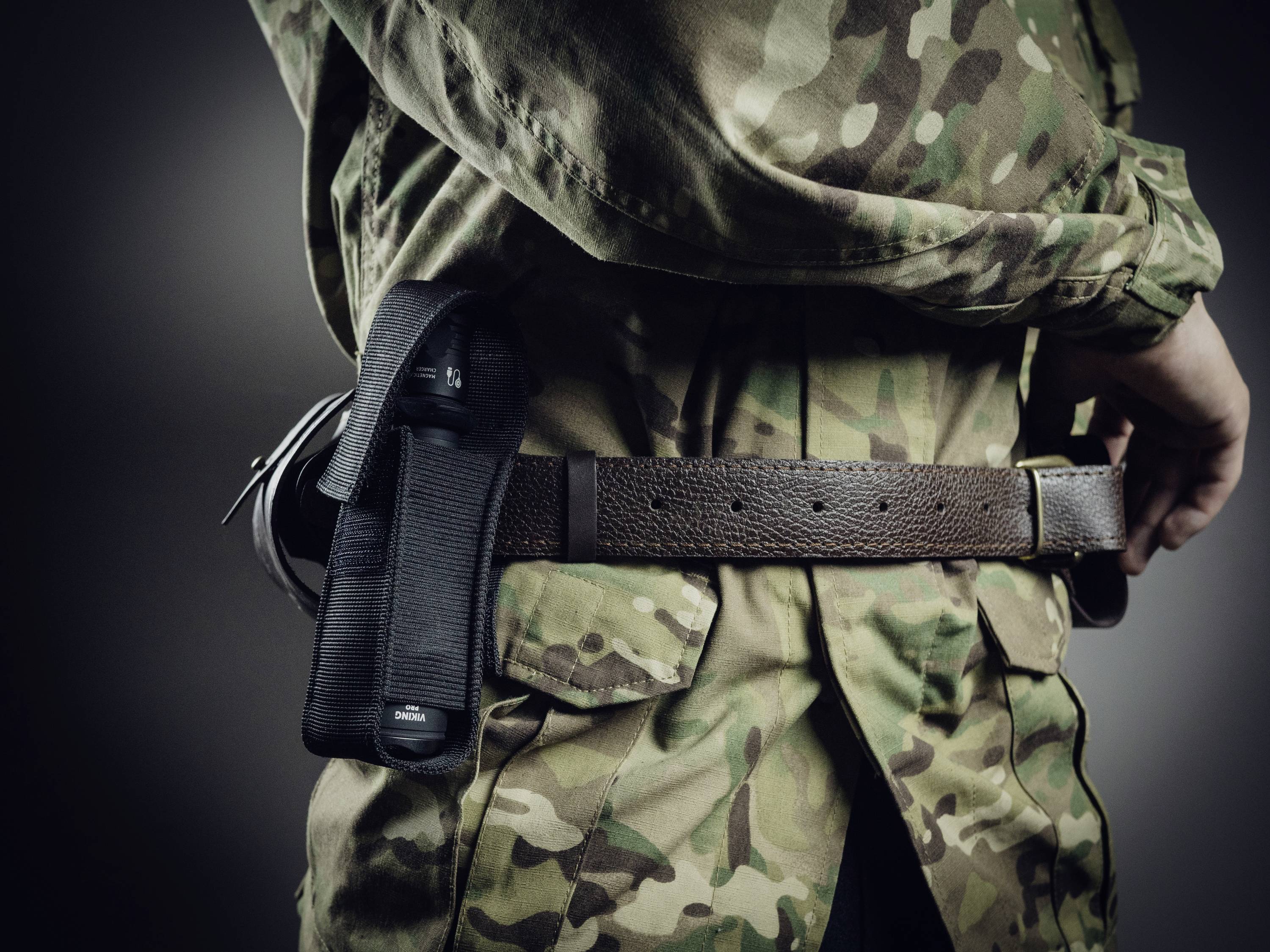 Eine Person in Tarnuniform trägt eine Handfeuerwaffe in einem Holster an einer Koppel, seitlich von hinten aufgenommen.