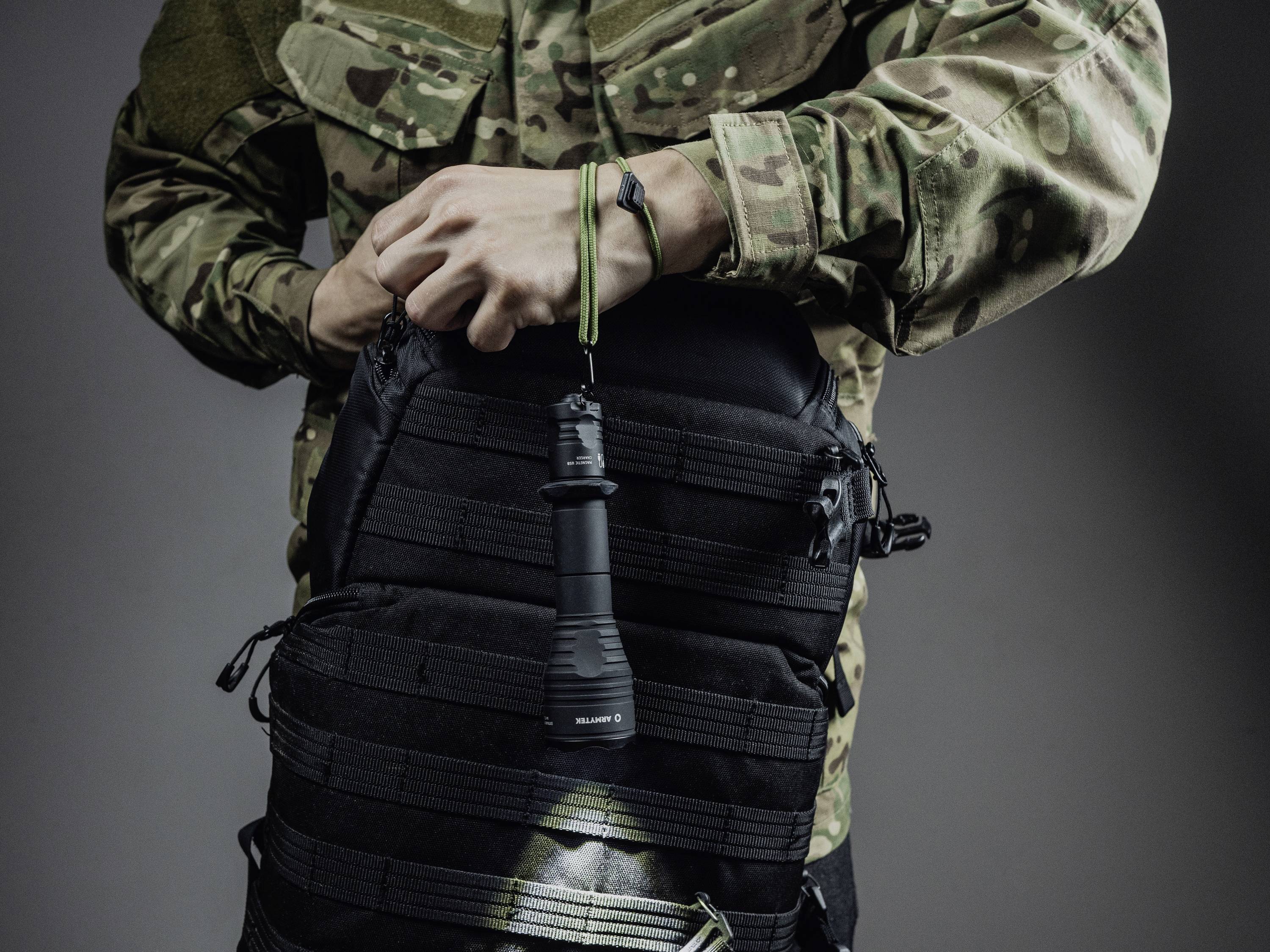 Ein Mensch in Tarnkleidung hält eine schwarze Taschenlampe, die an einem schwarzen Rucksack befestigt ist.