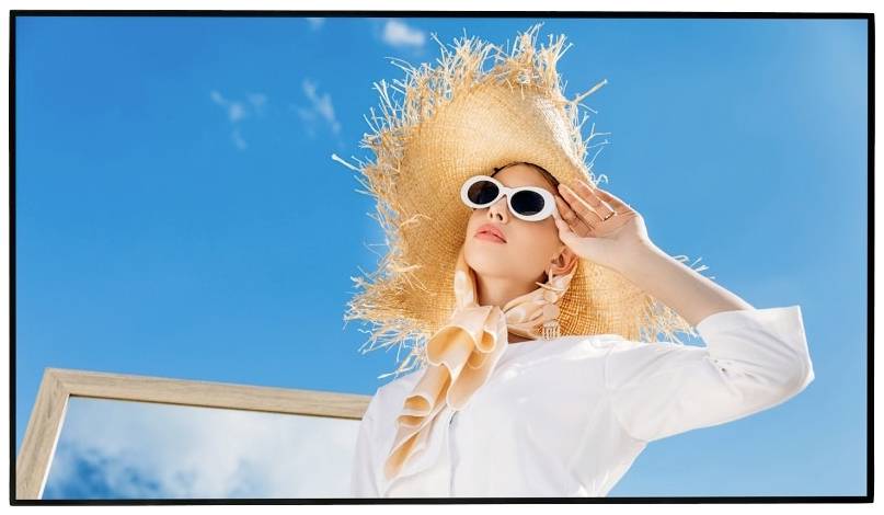 Frau mit Strohhut und Sonnenbrille posiert vor blauem Himmel, trägt ein weißes Hemd. Sie hält den Hut mit ihrer rechten Hand fest.