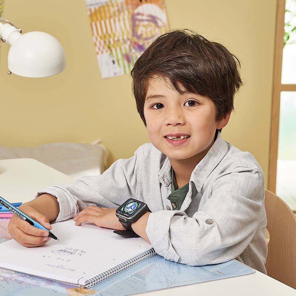 Ein Kind sitzt an einem Tisch und lächelt, während es auf ein Notizbuch schreibt. Es trägt eine Smartwatch.