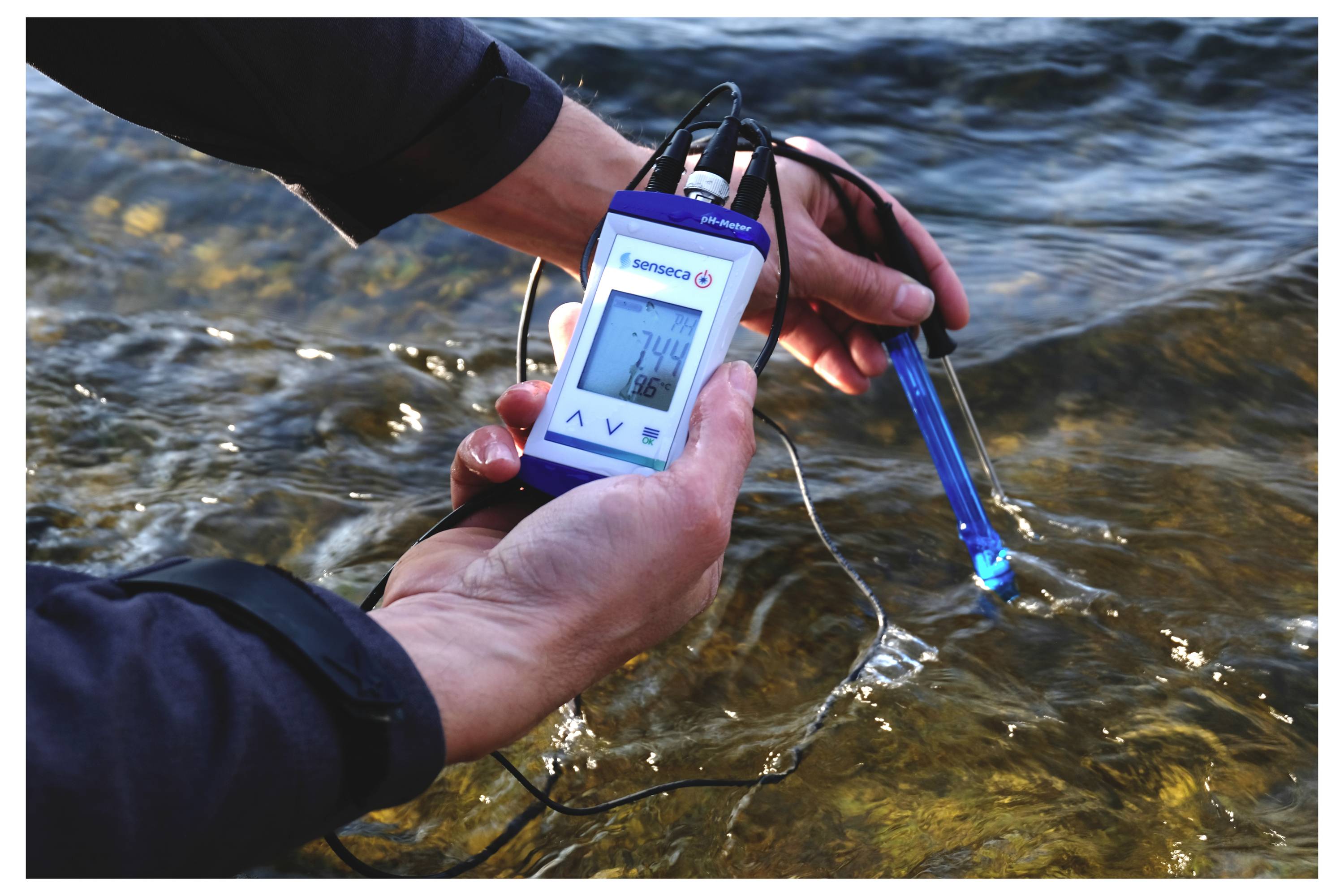 Eine Person, die ein digitales Gerät verwendet, um die Wasserqualität in einem Fluss zu messen, wobei eine Ablesung von 7,4 angezeigt wird.