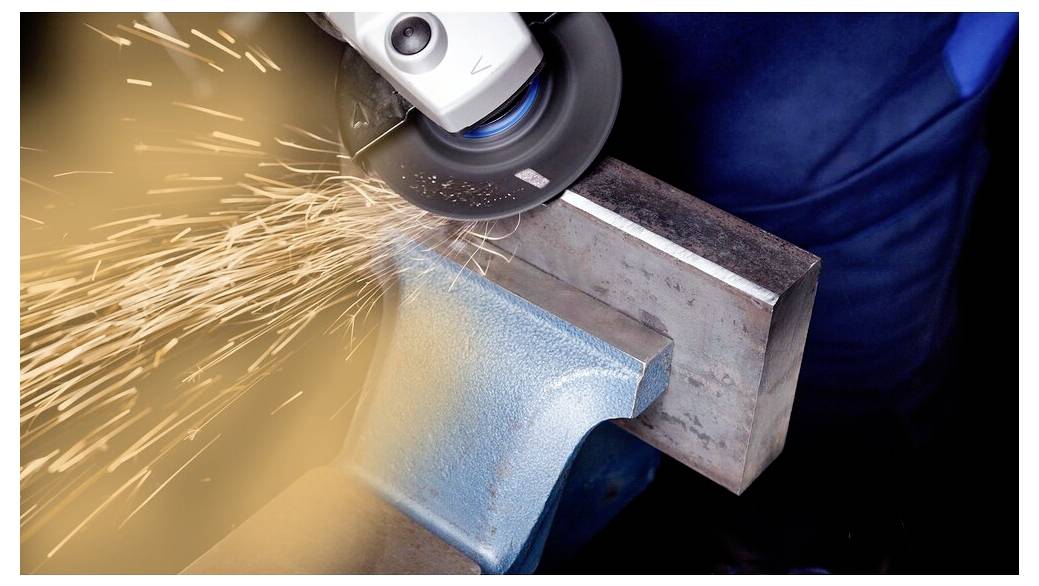 Ein Arbeiter verwendet einen Winkelschleifer, um Metall zu schneiden und erzeugt dabei helle Funken. Das Metall ist in einem blauen Schraubstock befestigt.