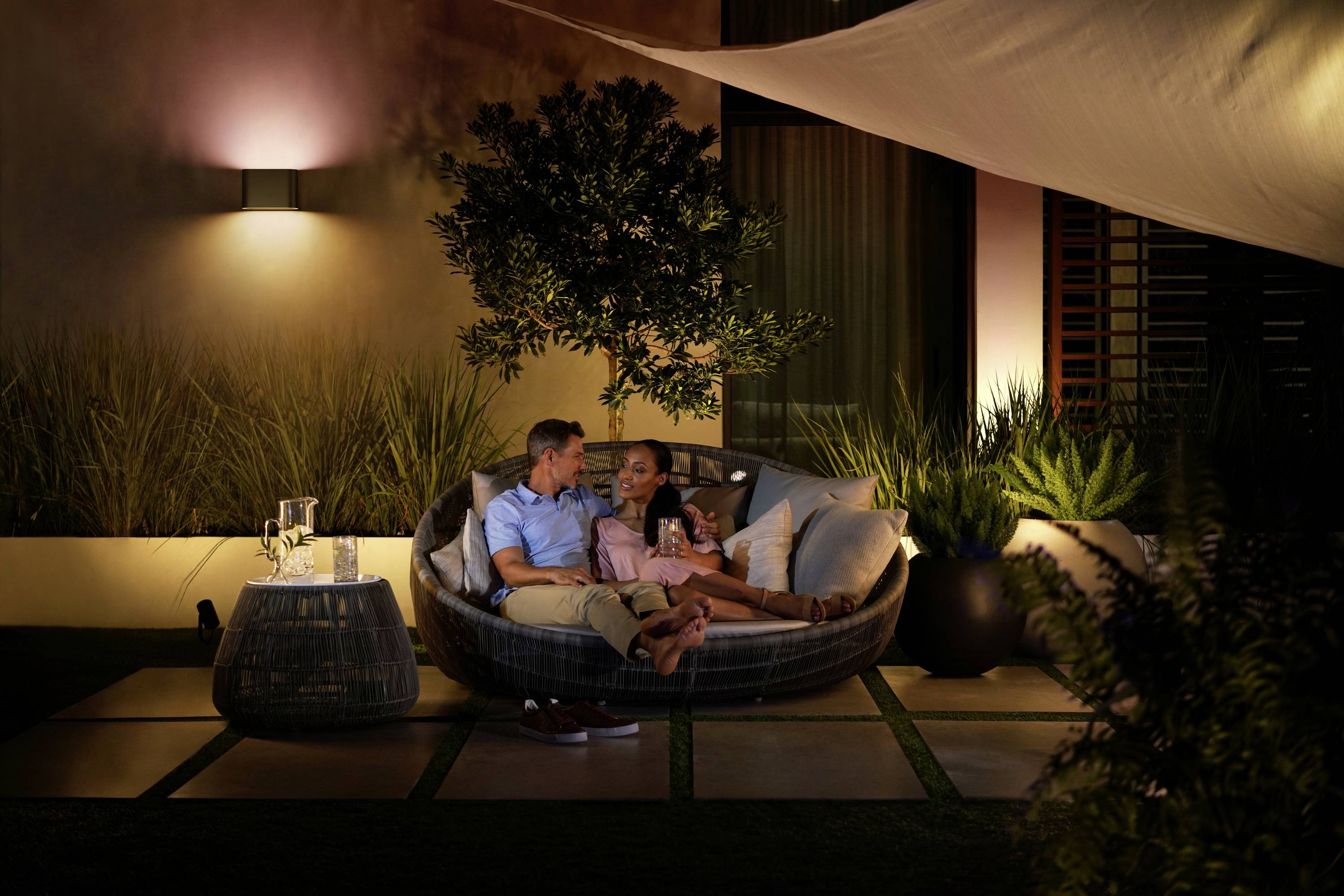 Ein Paar sitzt entspannt in einem runden Loungemöbel im Garten, umgeben von Pflanzen und gedämpftem Licht, mit Getränken auf einem Beistelltisch.