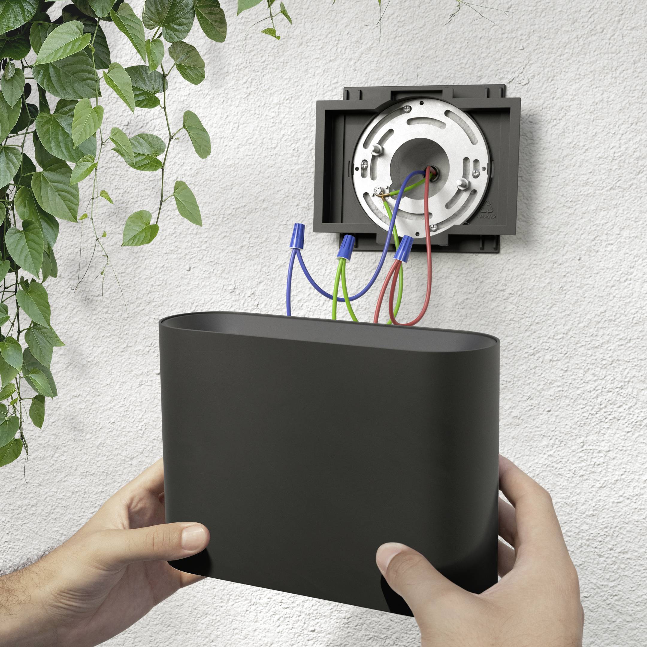 Zwei Hände halten ein schwarzes Gerät unter einer offenen Steckdose mit sichtbaren blauen und roten Kabeln an einer weißen Wand.