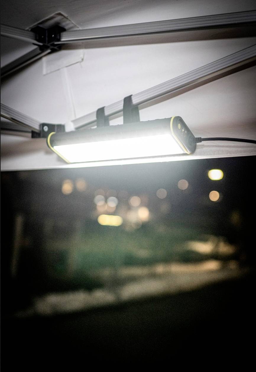 Helles LED-Licht, das an einer Decke montiert ist, beleuchtet einen nächtlichen Außenbereich. Unscharfer Hintergrund mit Bäumen und Lichtern.