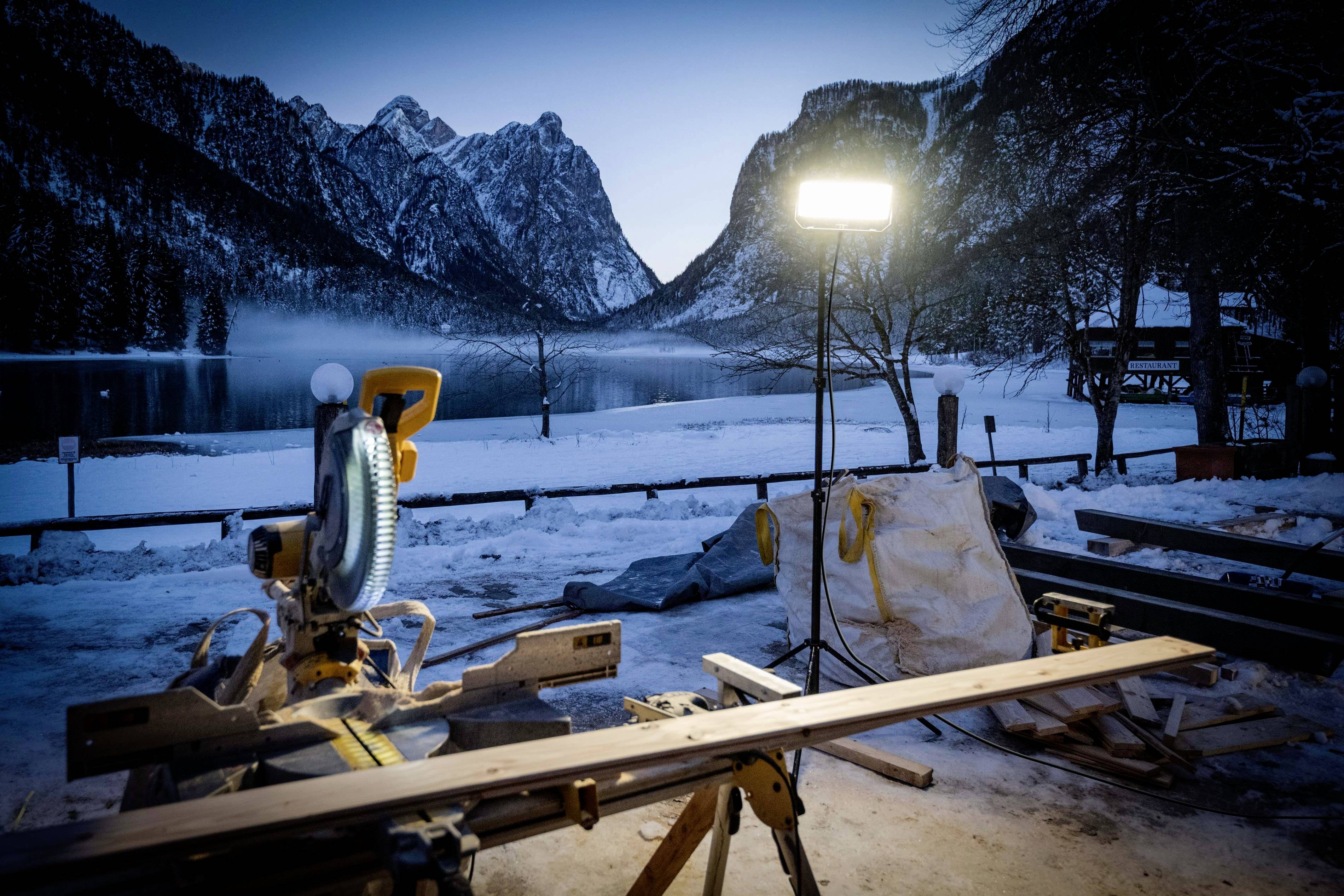 Holzsäge auf Tisch vor verschneitem Bergsee bei Dämmerung, beleuchtet von hellem Baustrahler. Majestätische Berglandschaft im Hintergrund.