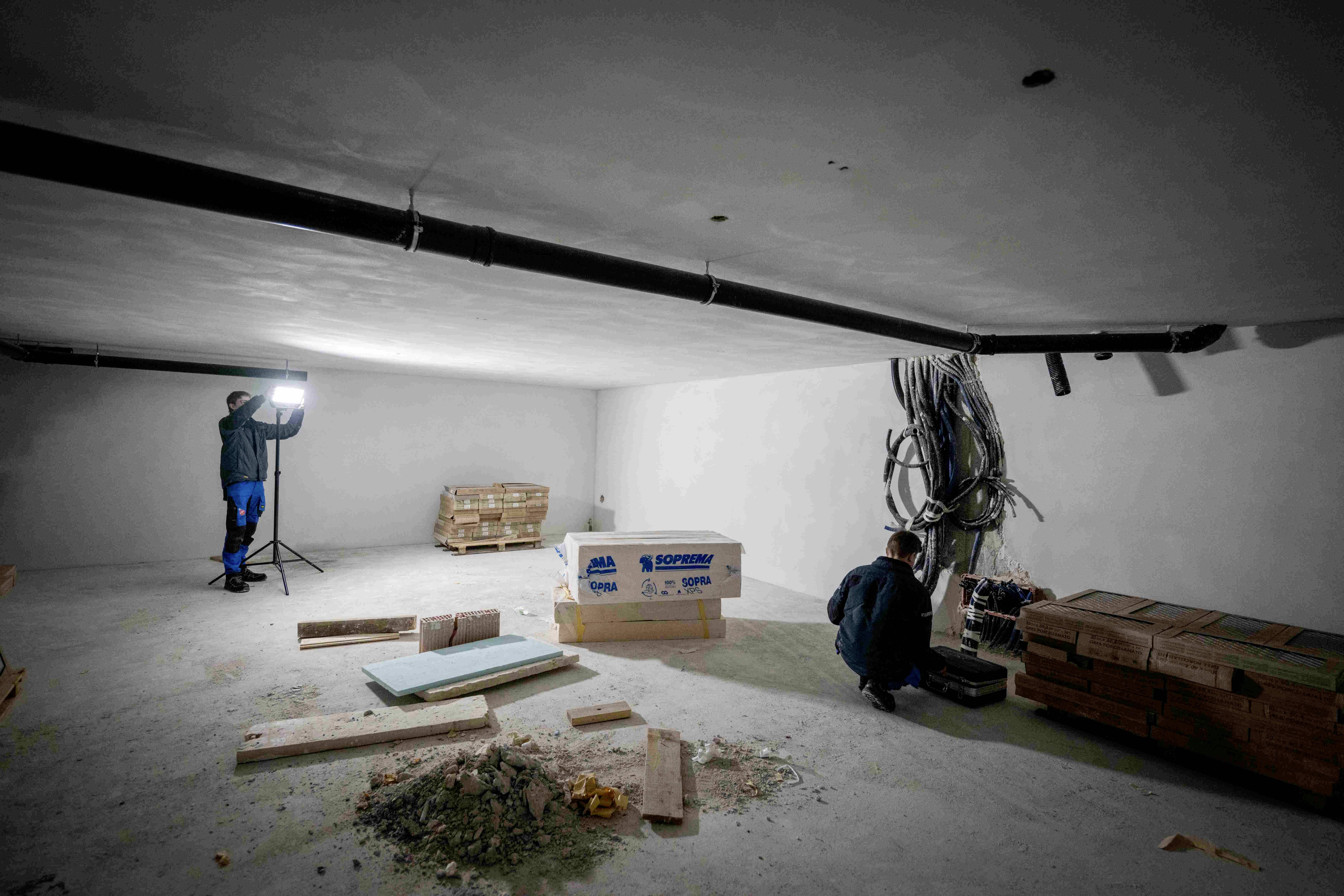 Eine leere Baustelle in einem Keller, zwei Arbeiter, Materialien und Werkzeuge verteilt. Einer der Arbeiter steht vor einer Lampe.