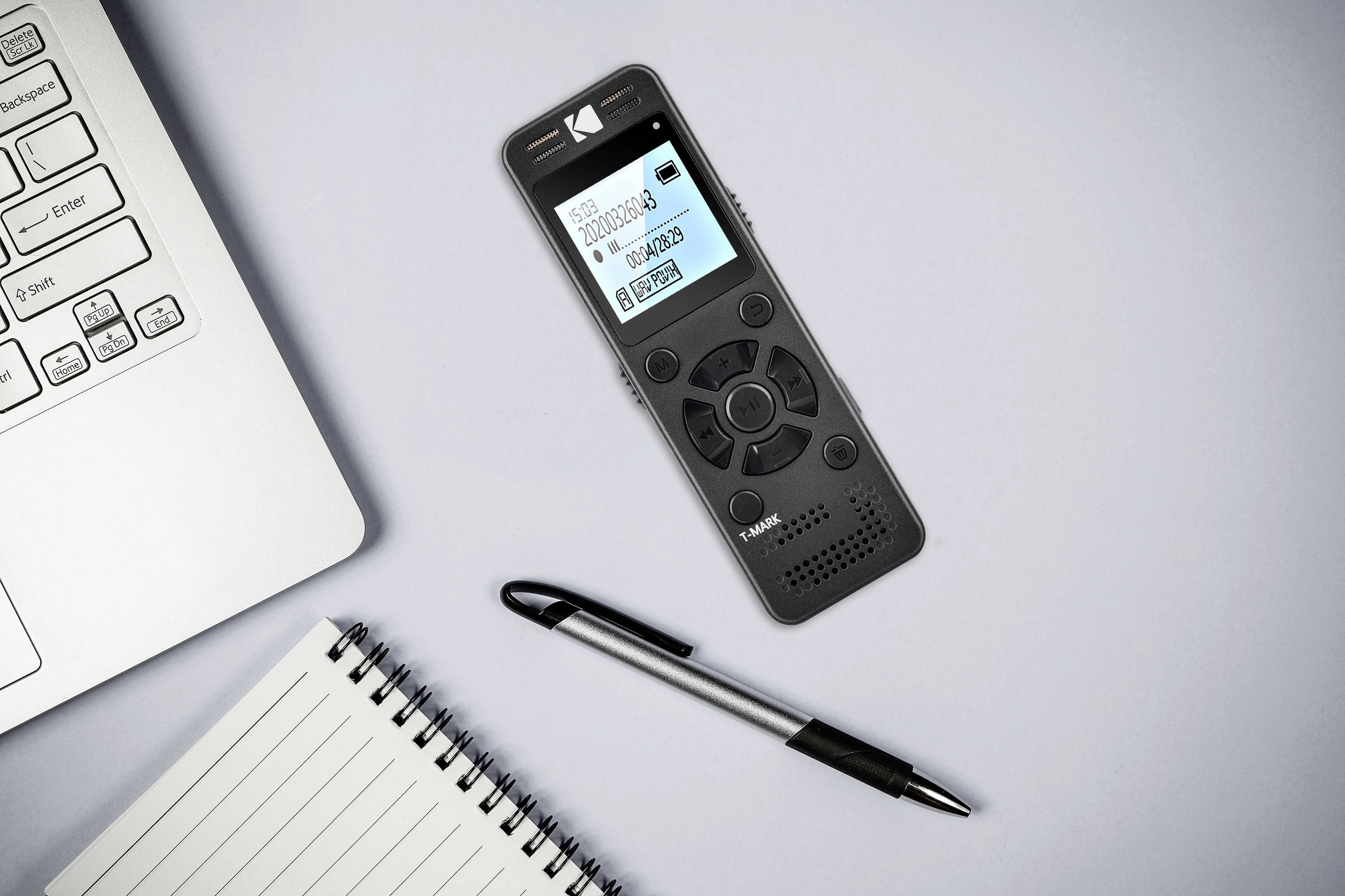 Diktiergerät mit digitalem Display liegt neben Laptop, Stift und Notizblock auf einem Tisch. Gerät zeigt Aufnahmeinformationen an.