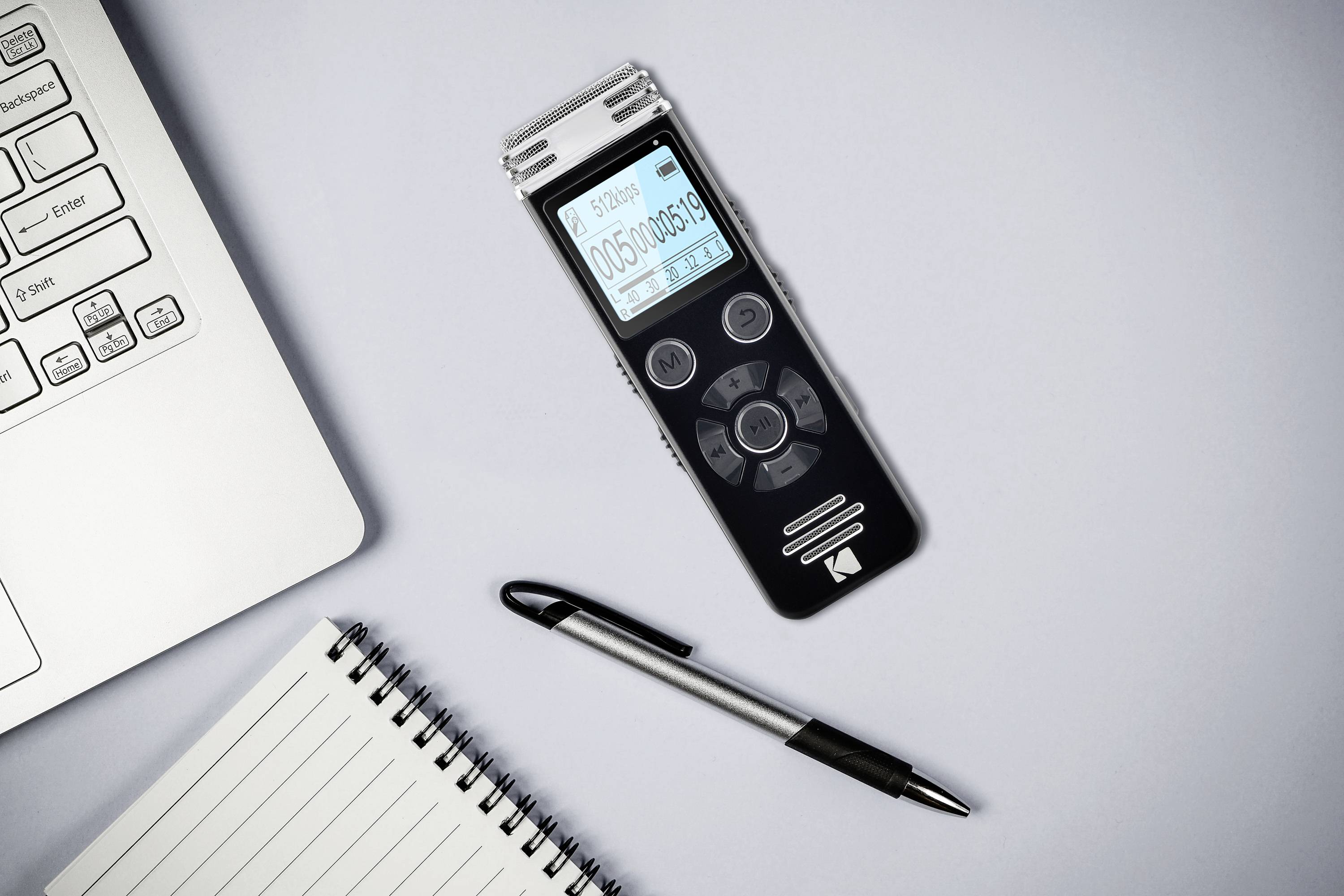 Diktiergerät mit Display zeigt Aufnahmezeit; liegt neben einem Laptop, einem Notizblock und einem Stift auf einem grauen Tisch.