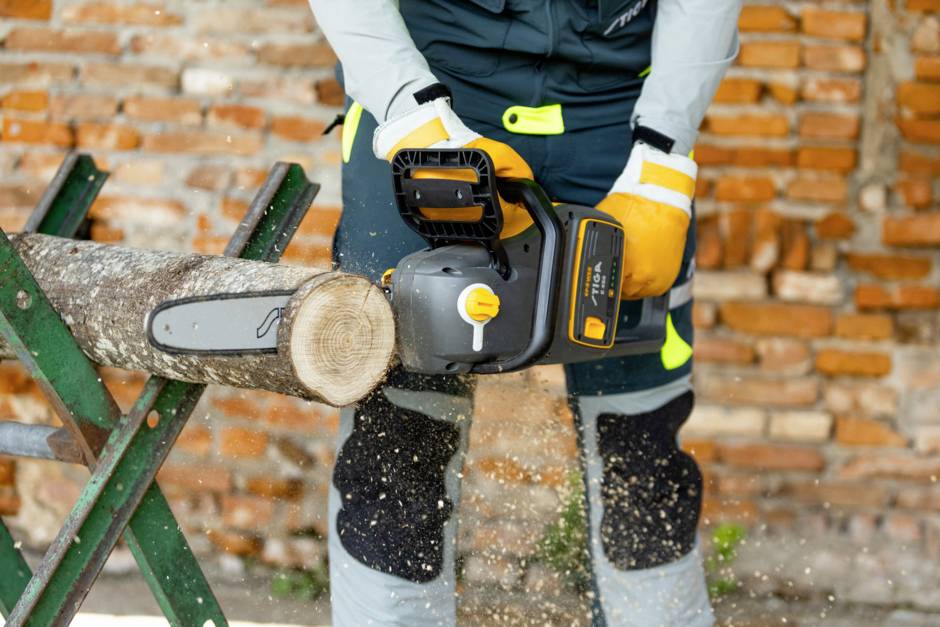 Eine Person sägt mit einer elektrischen Kettensäge einen Holzstamm, getragen von einer grünen Halterung. Ziegelwand im Hintergrund.