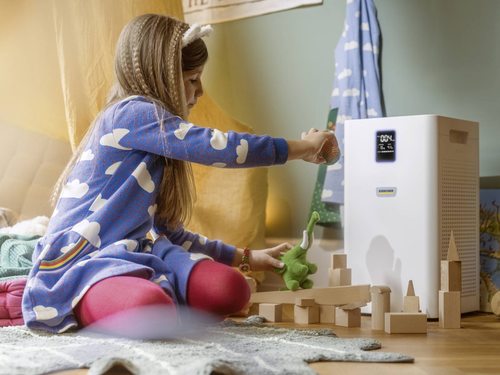 Mädchen in blauem Kleid spielt mit Holzblöcken neben einem Luftreiniger im Kinderzimmer. Luftreiniger zeigt '02%' auf Display.