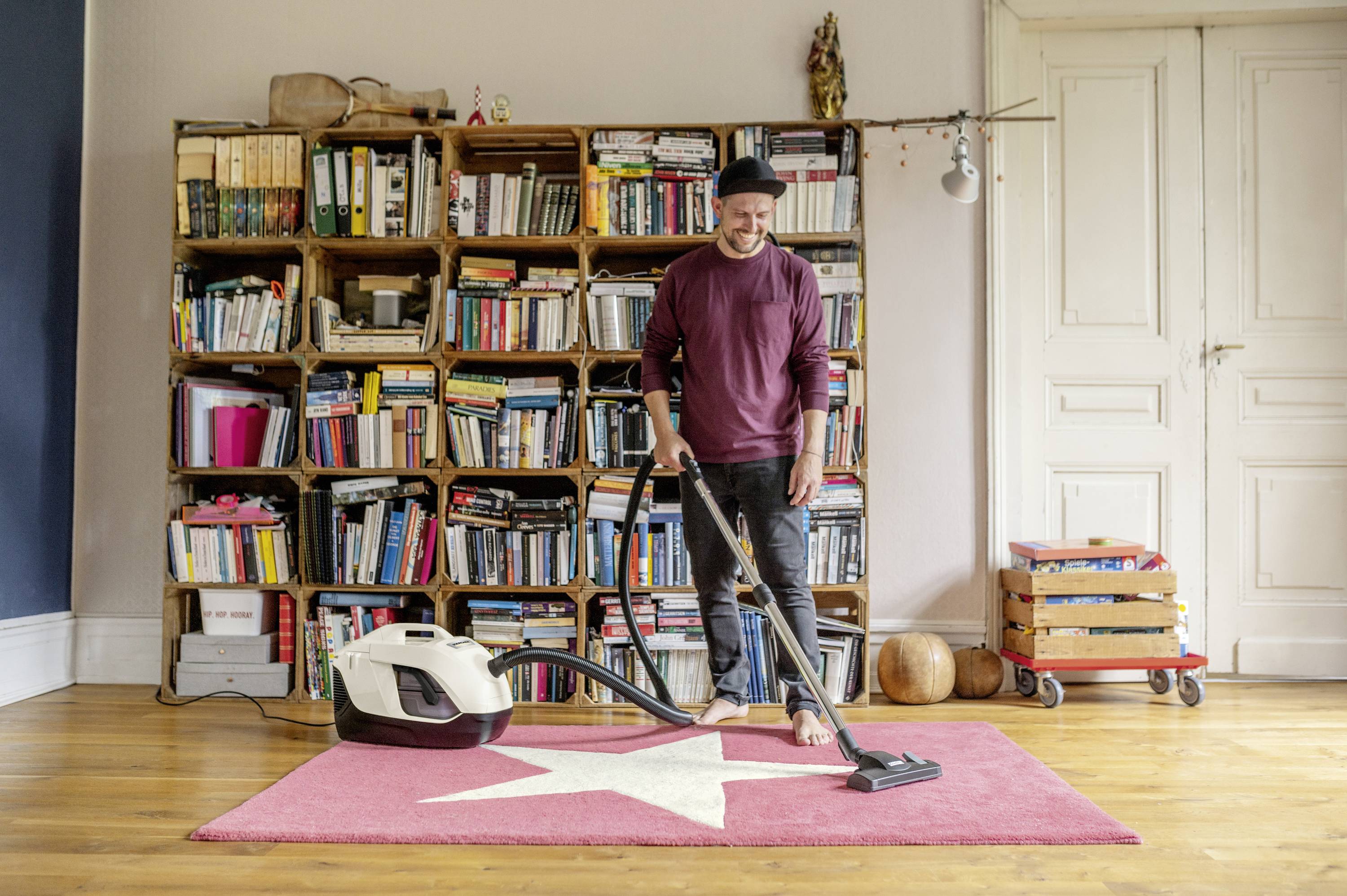 Ein Mann saugt einen rosafarbenen Teppich mit einem Sternmotiv in einem Wohnzimmer mit Bücherregalen ab.